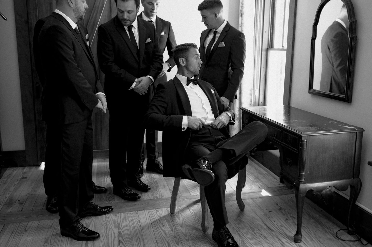 Black and white photo of groom seated at desk surrounded by groomsmen in tuxedos.