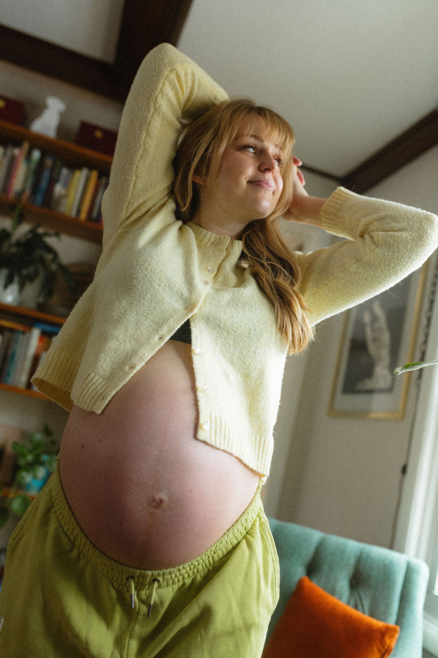Pregnant woman in a yellow crop sweater and green pants posing indoors with hands behind her head.