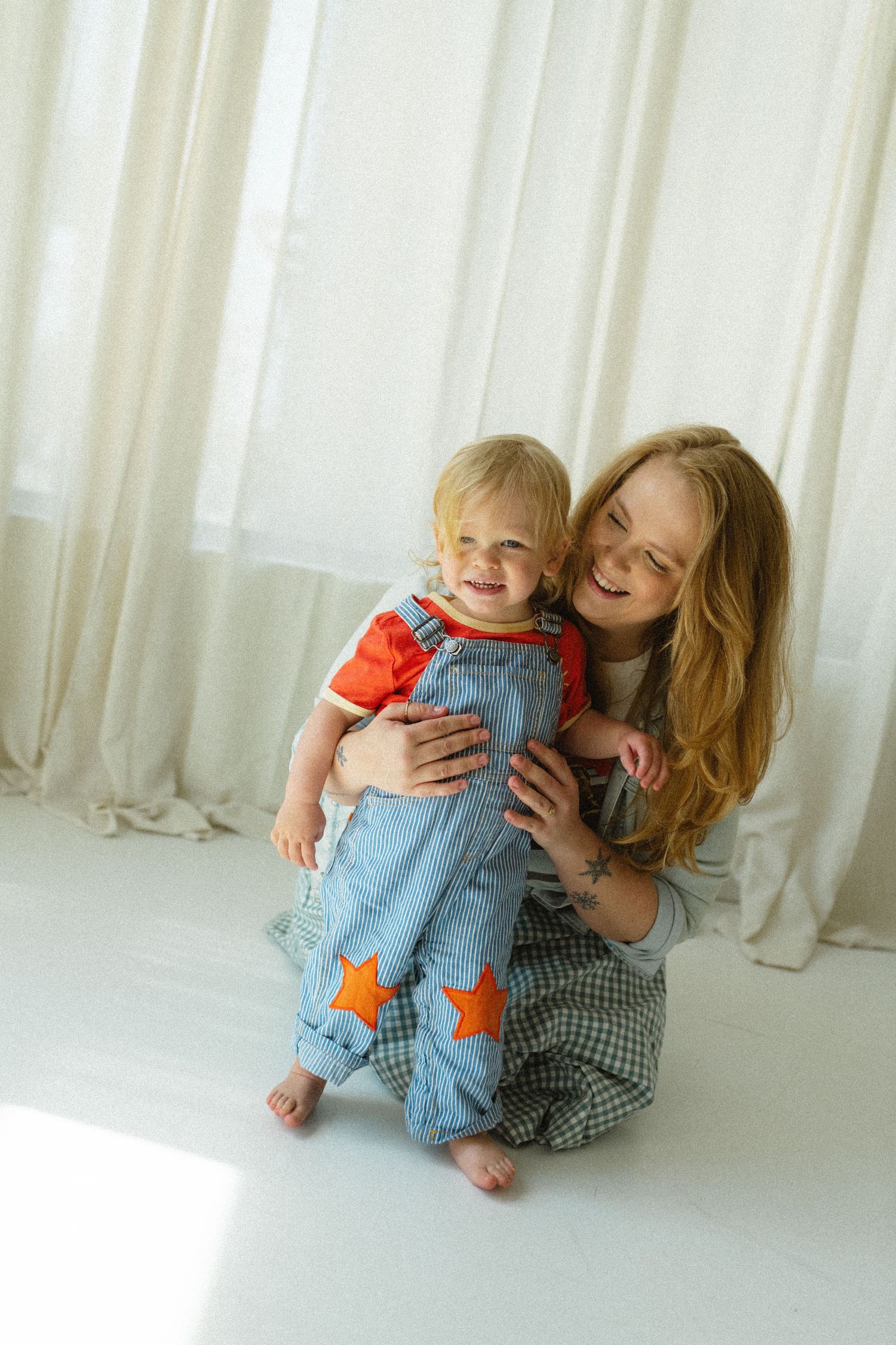 Smiling mother hugging her happy blonde toddler in denim star overalls indoors.