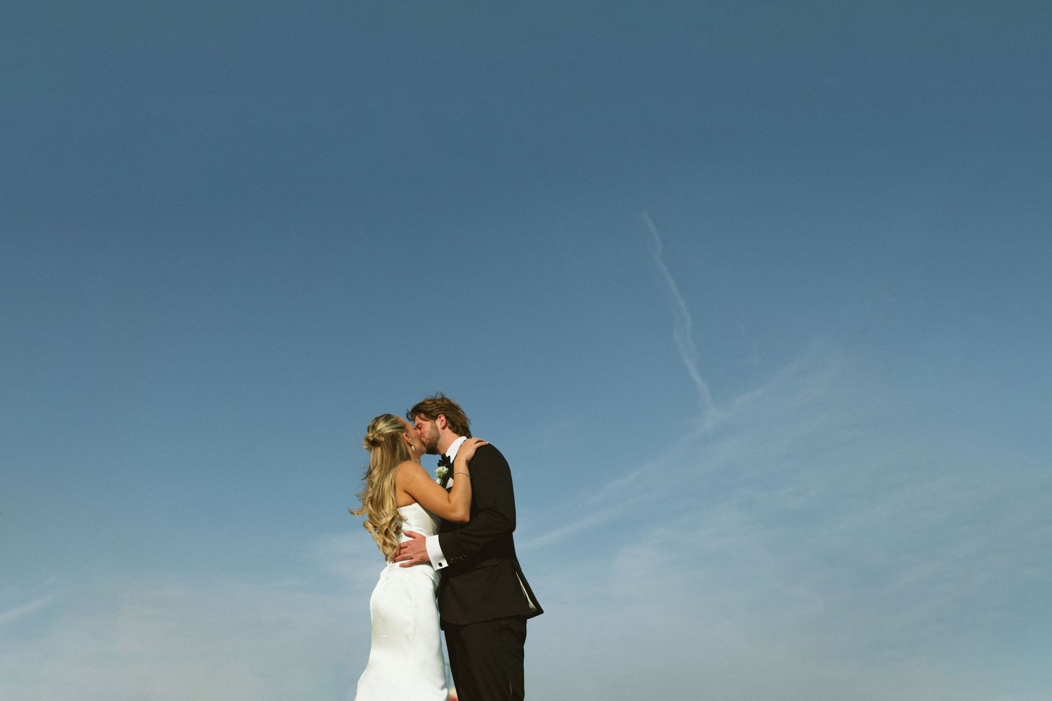 Bride and groom share romantic kiss against clear blue sky backdrop.
