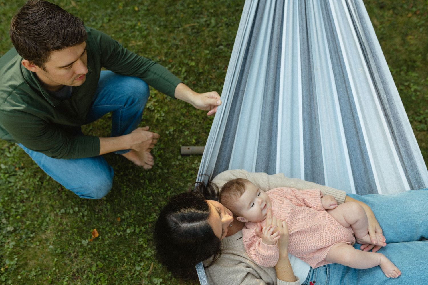 Father kneels beside mother and baby resting in a striped hammock in a lush green garden.