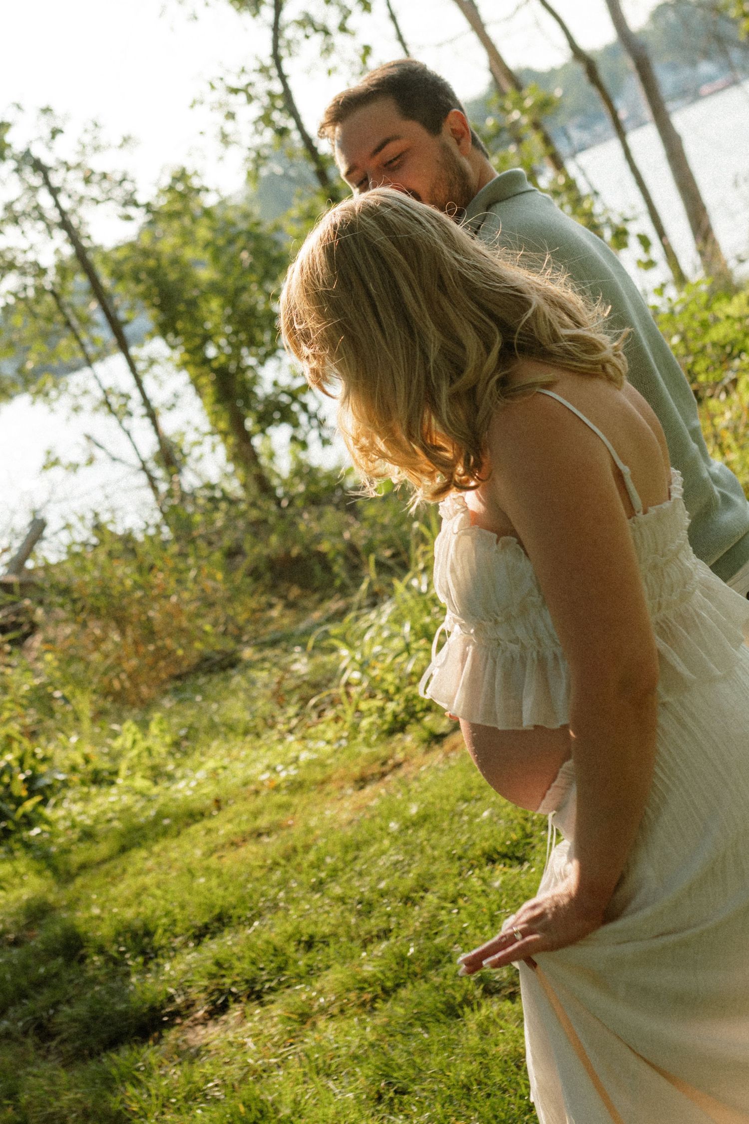A man kissing a pregnant woman on the head while embracing her outdoors near a sunlit lake.