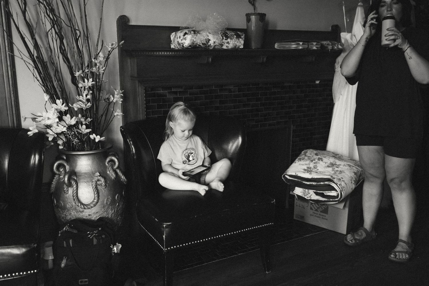 Black and white photo of child sitting on ottoman by fireplace.