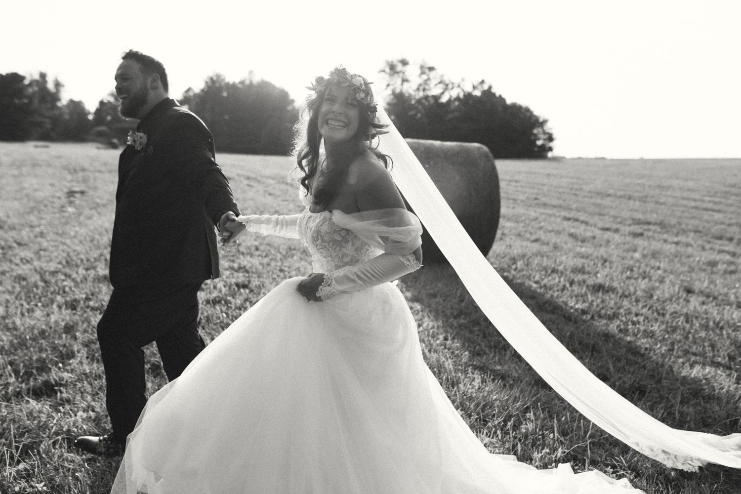 Black and white photo of bride and groom walking through farm field with flowing veil.