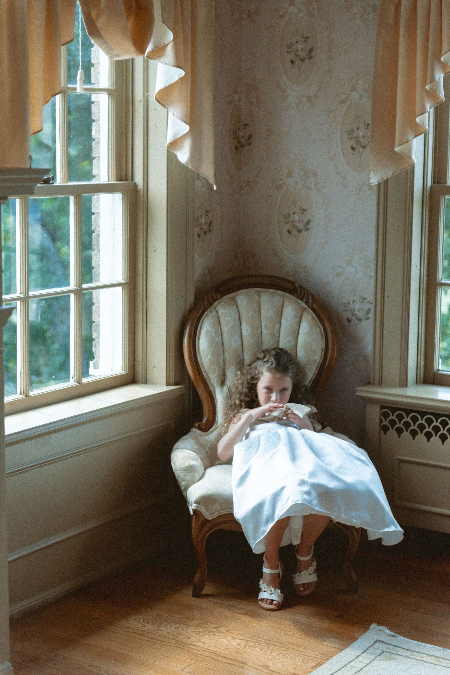 Adorable curly-haired flower girl in white dress sitting in antique chair by bright windows.