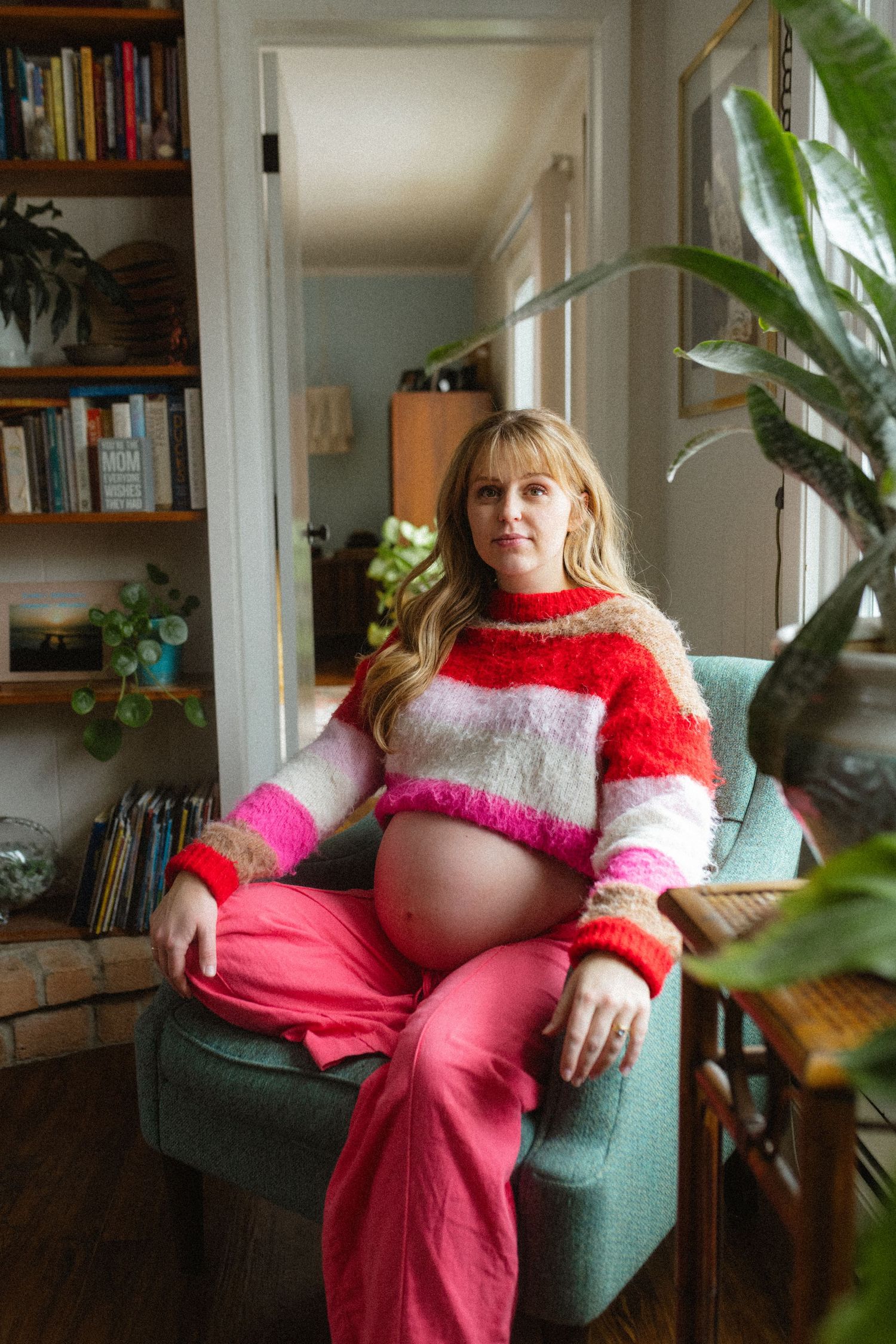 Pregnant woman in a colorful striped sweater sitting in an armchair surrounded by plants and bookshelves.