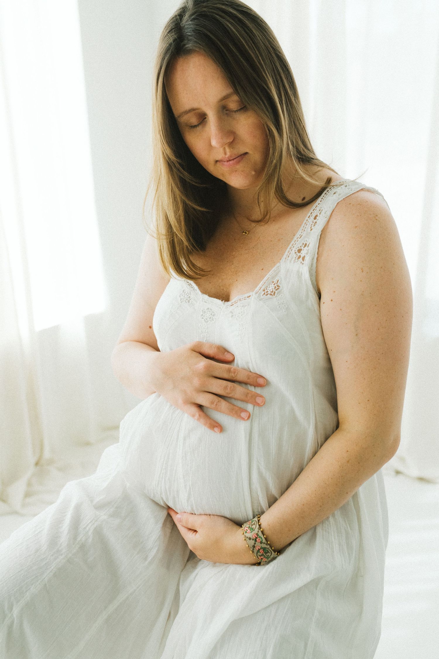 Serene pregnant woman in white lace dress cradling baby bump in softly lit studio setting.