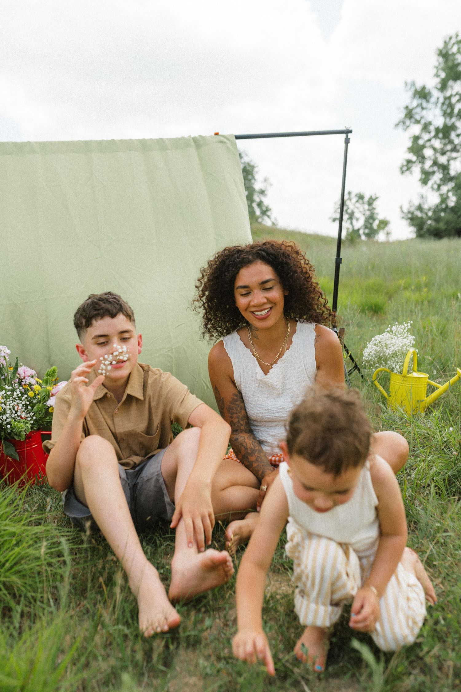 Curly-haired mother smiles as son blows dandelion and toddler plays on grass outdoors.