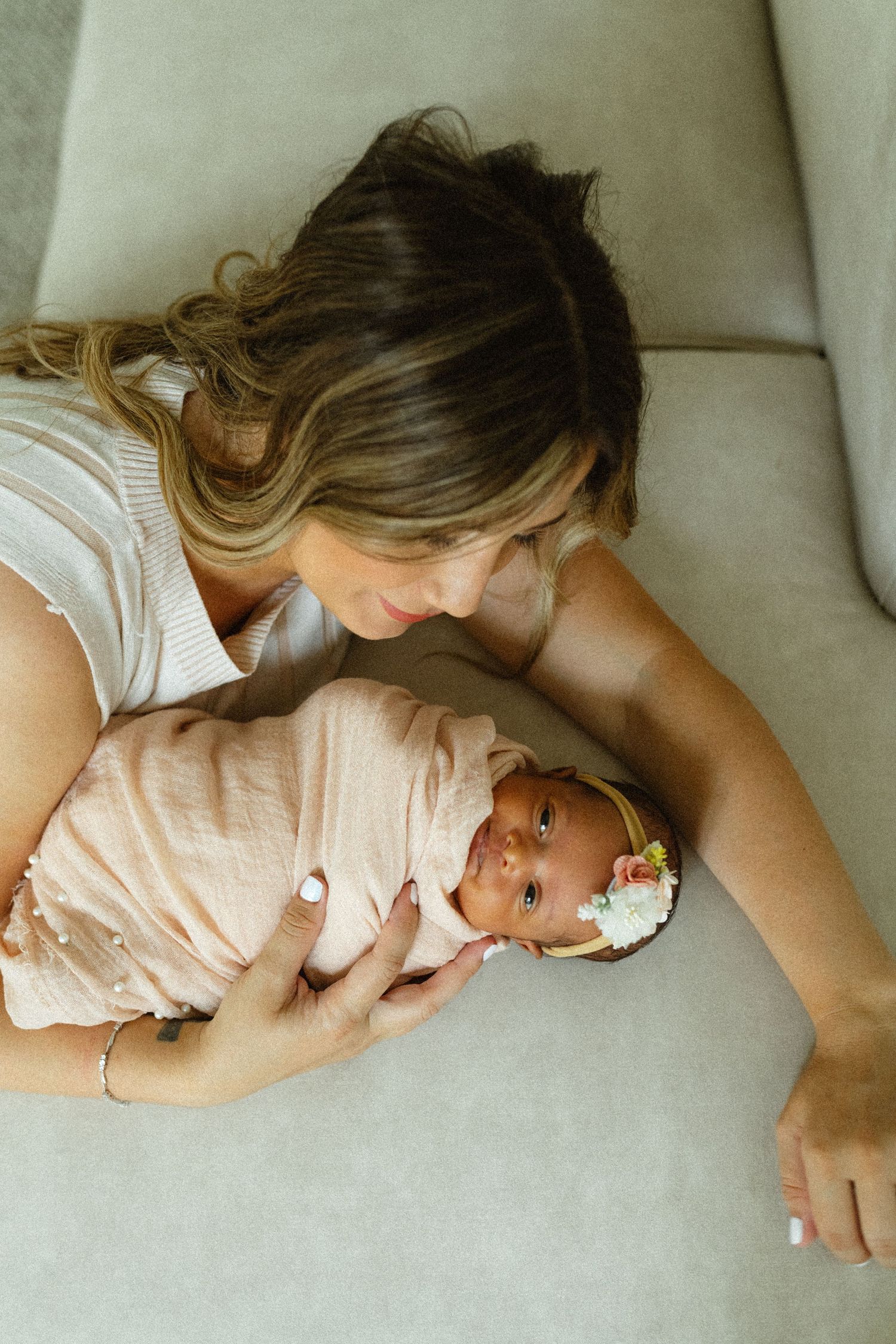 Mother gazing lovingly at swaddled newborn baby with floral headband lying on gray couch.