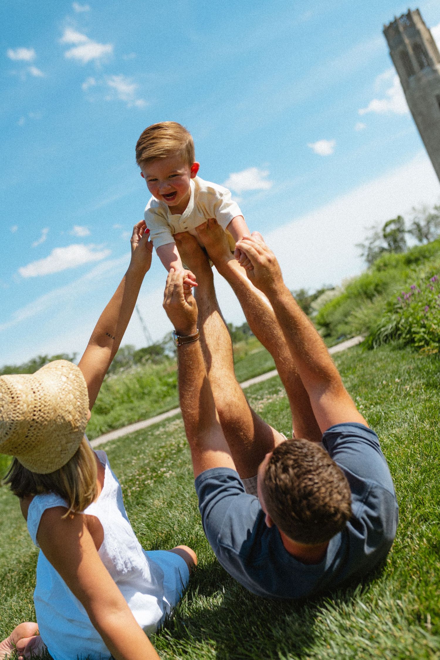 Family sitting on green grass as father lifts giggling toddler into the blue sky outdoors.