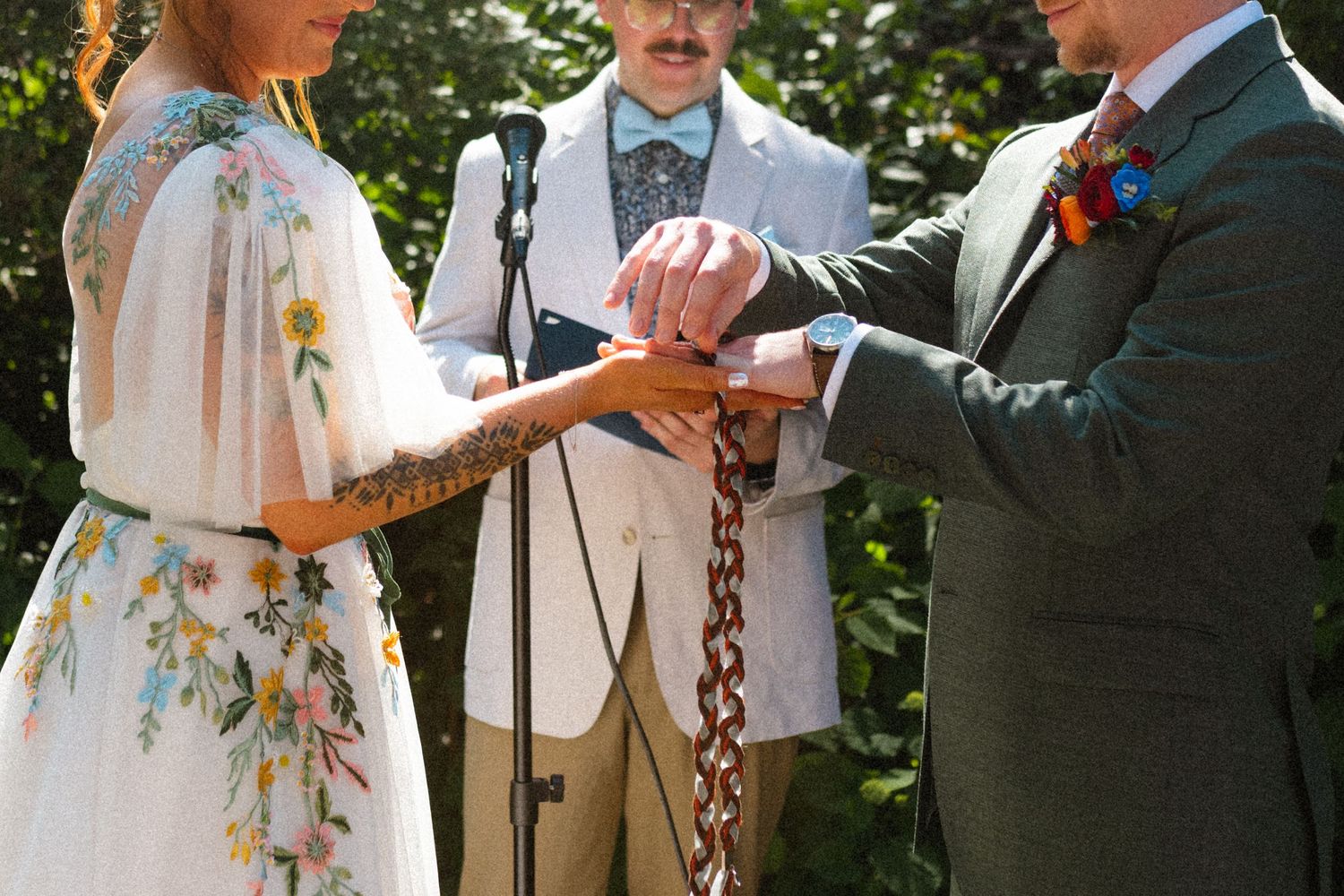 Bride and groom performing handfasting ceremony with chain, officiant standing behind them outdoors.