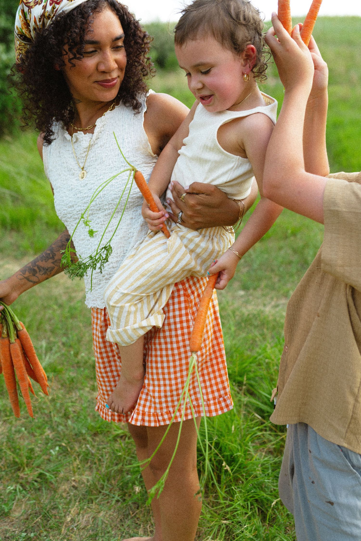 Smiling mother holds toddler while boy waves fresh carrots during outdoor family photo session.