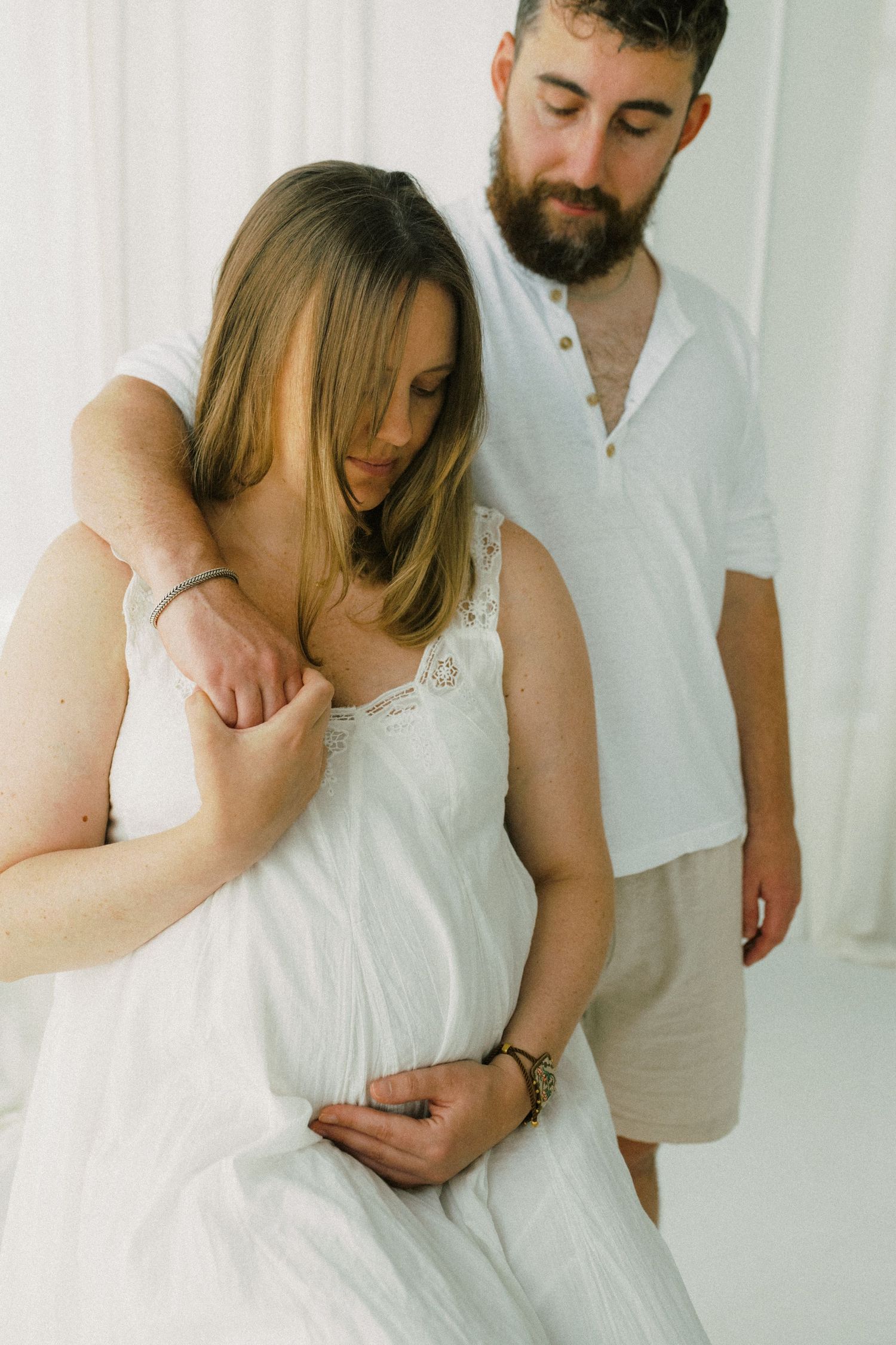 Couple sharing tender moment during maternity photoshoot, man embracing pregnant woman in white dress.