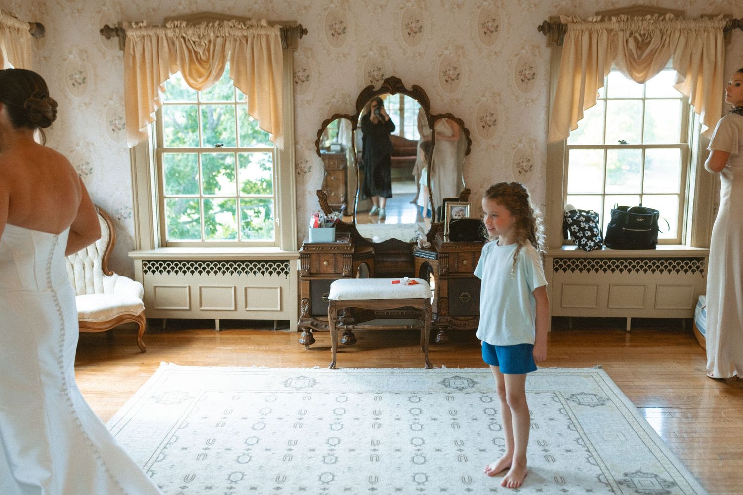 Young girl in blue shorts watching bride in white gown in vintage Victorian dressing room.