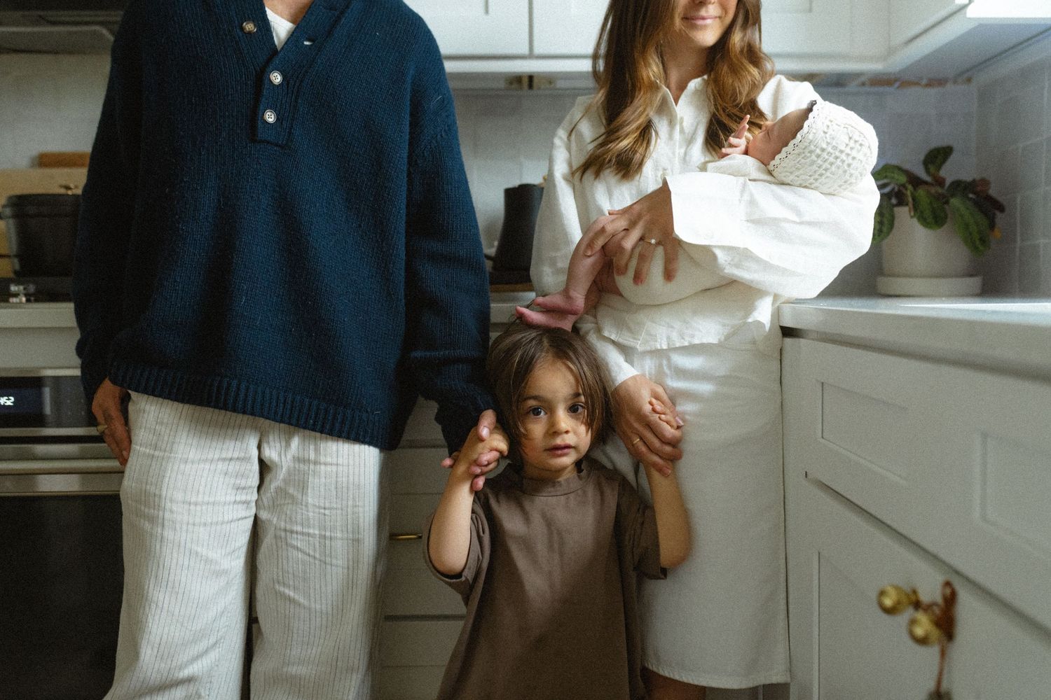 Family of four with newborn and toddler standing together in a bright white kitchen.