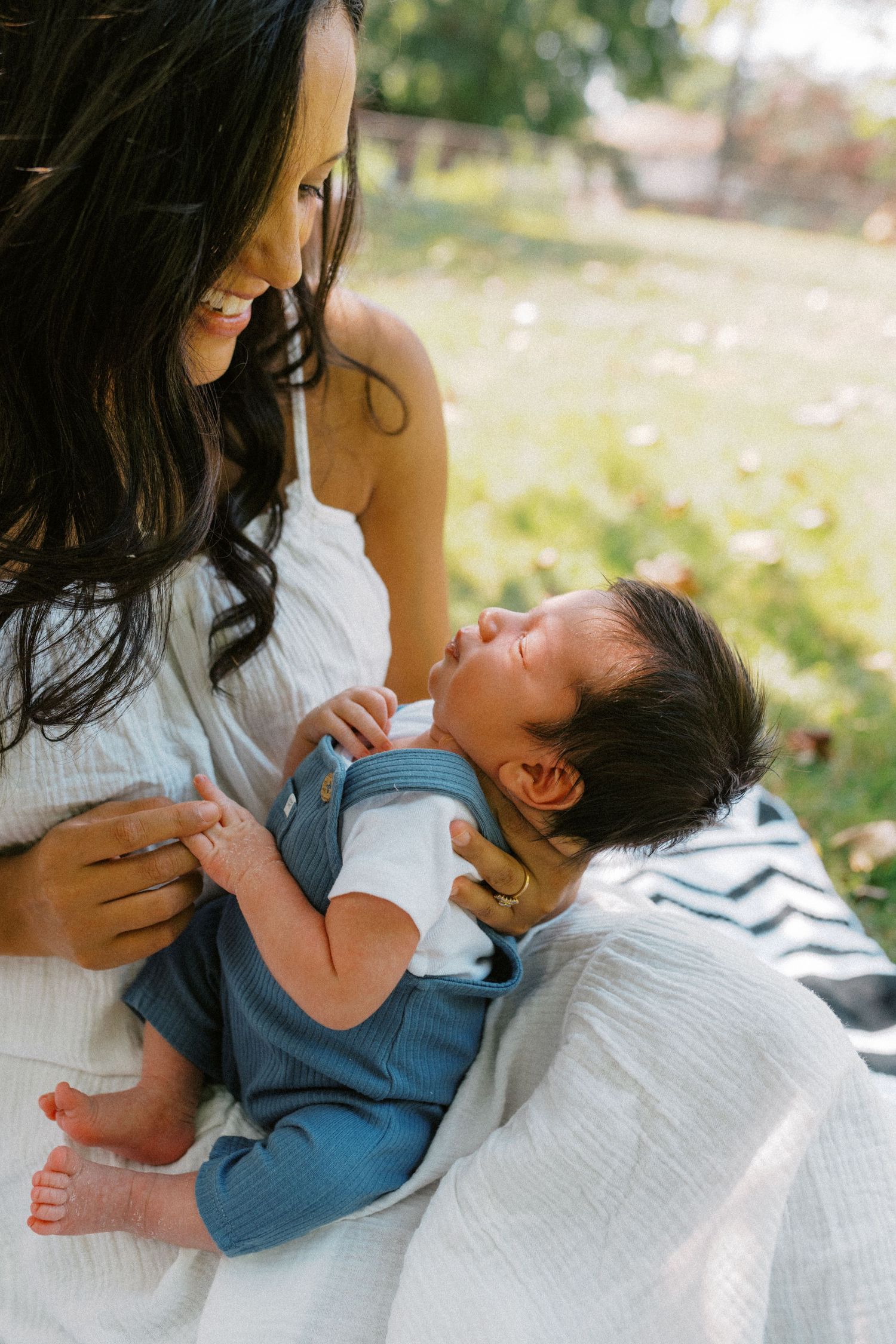 Smiling mother in white dress cradling her newborn baby outdoors in soft natural light.