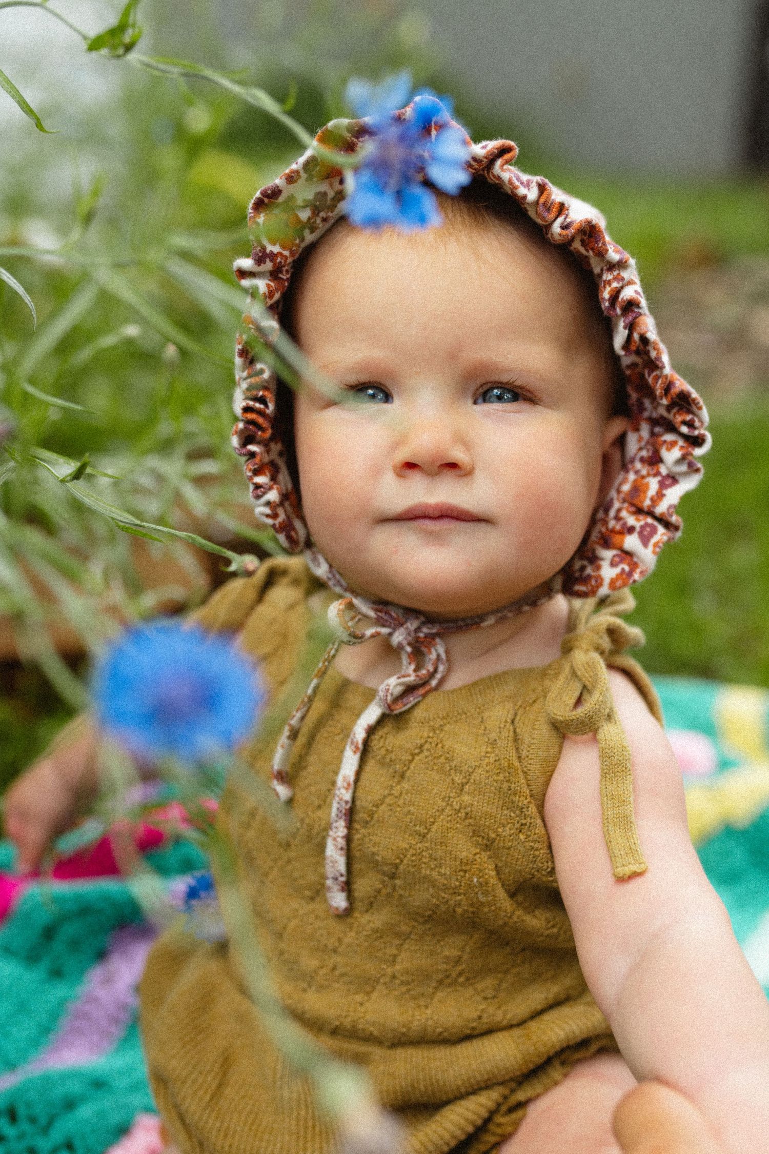 Adorable baby in a floral bonnet and olive romper surrounded by blue wildflowers outdoors.