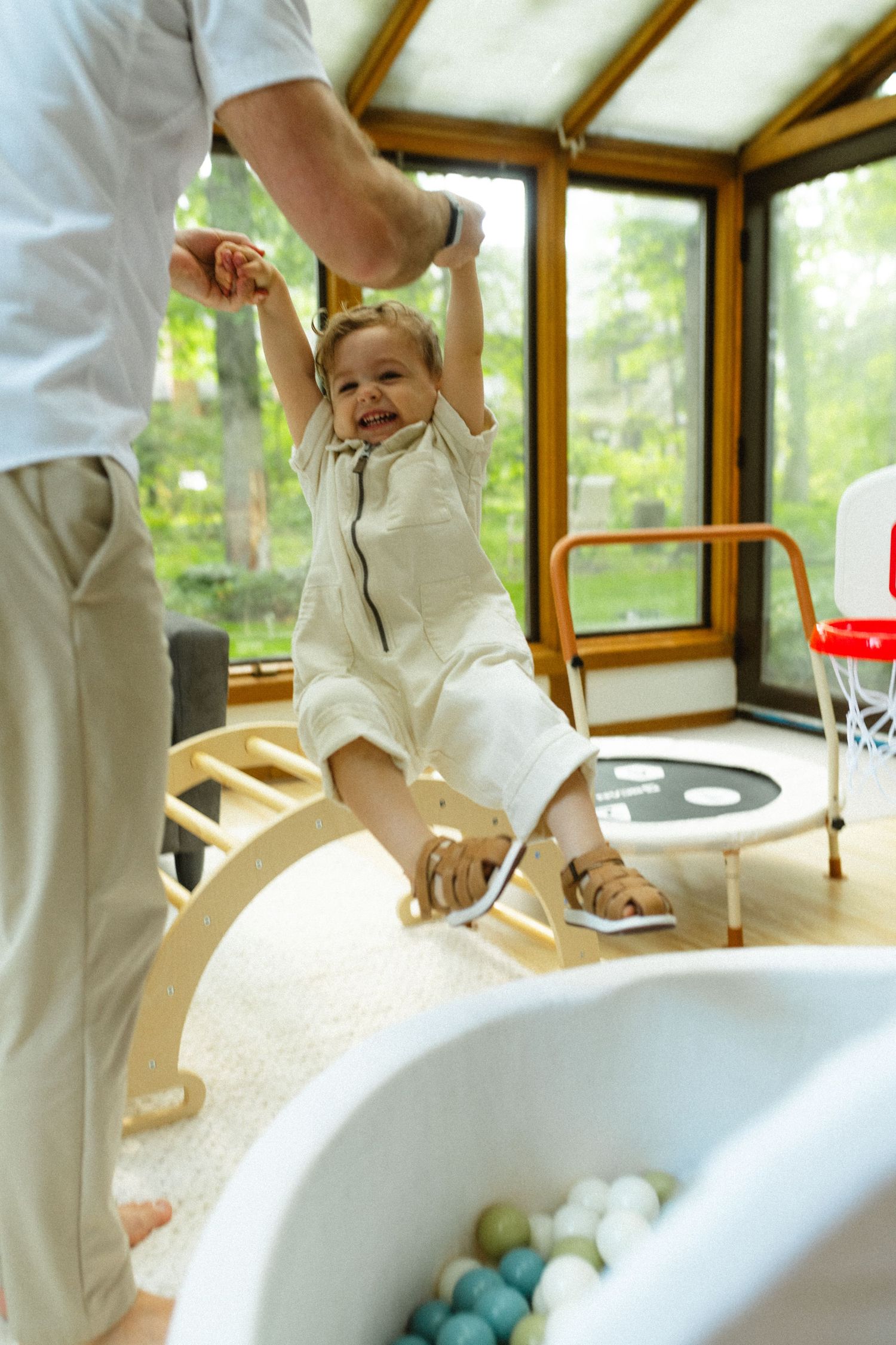 Father helping laughing toddler balance on wooden rocker board in bright sunroom playroom.