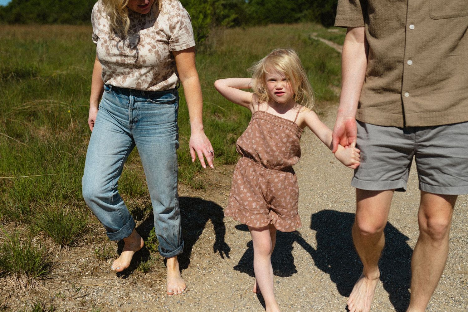 Young blonde girl holds parents hands walking barefoot on sandy path on sunny day.