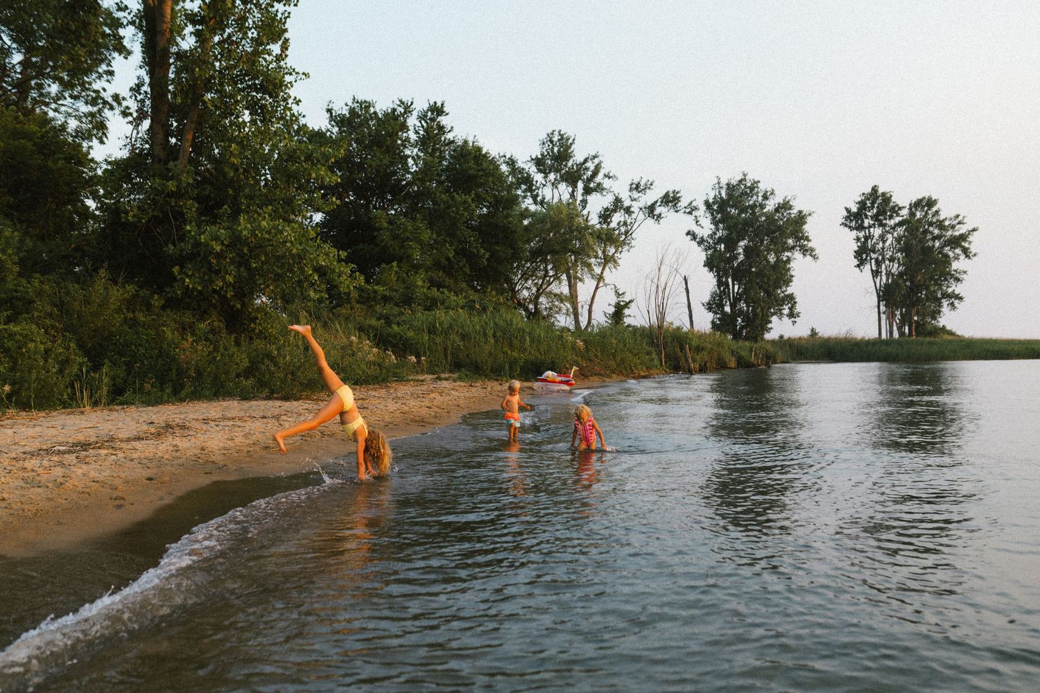 Children playing and doing cartwheels on a sandy beach shoreline surrounded by lush green trees.