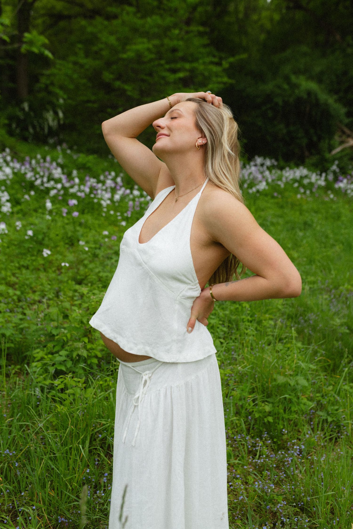 Pregnant woman in white linen outfit posing among wildflowers and lush green foliage.