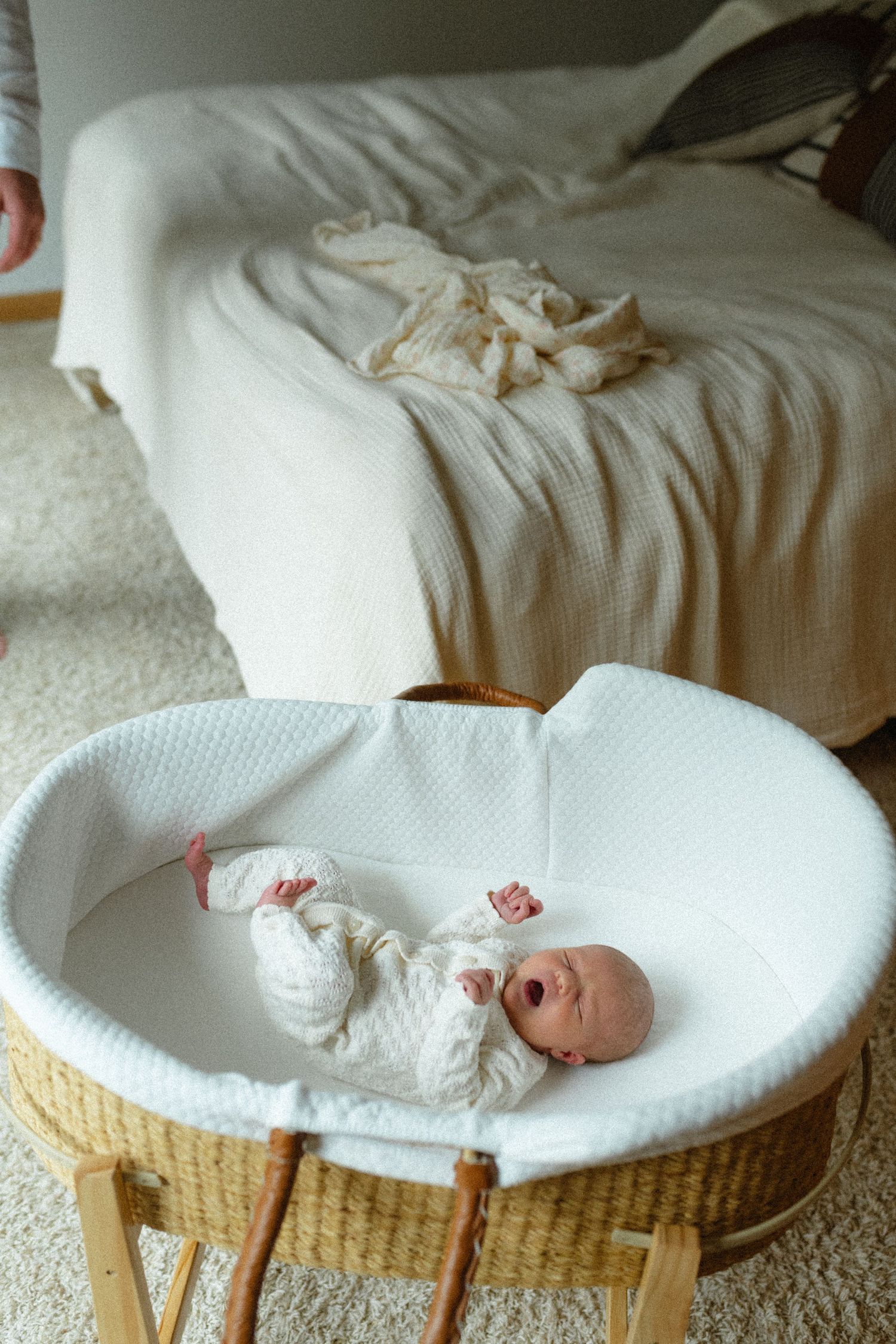 Newborn baby in white onesie yawning in wicker Moses basket beside bed.
