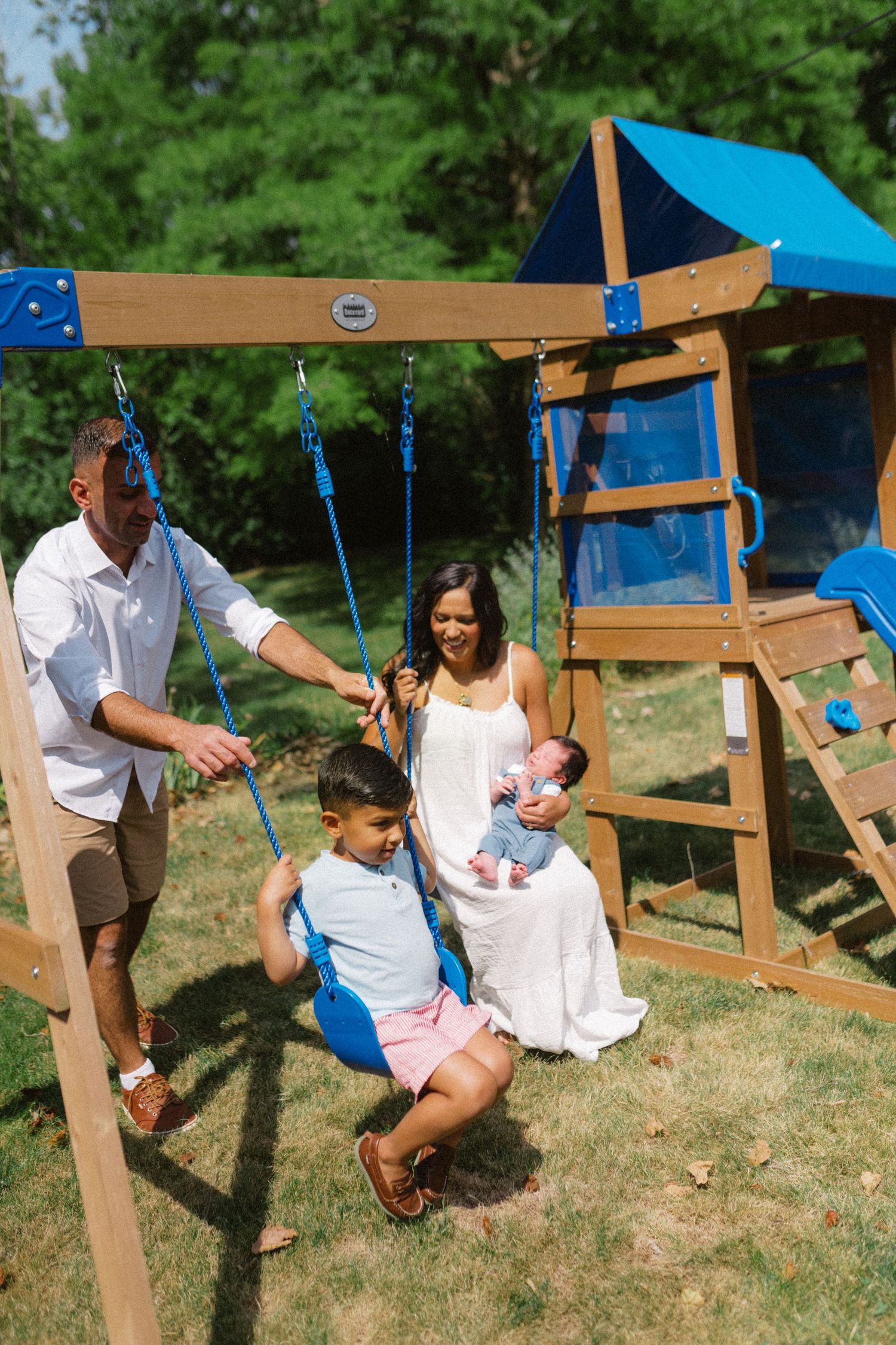 Family enjoying outdoor playtime at backyard swing set with mother holding newborn baby on sunny day.