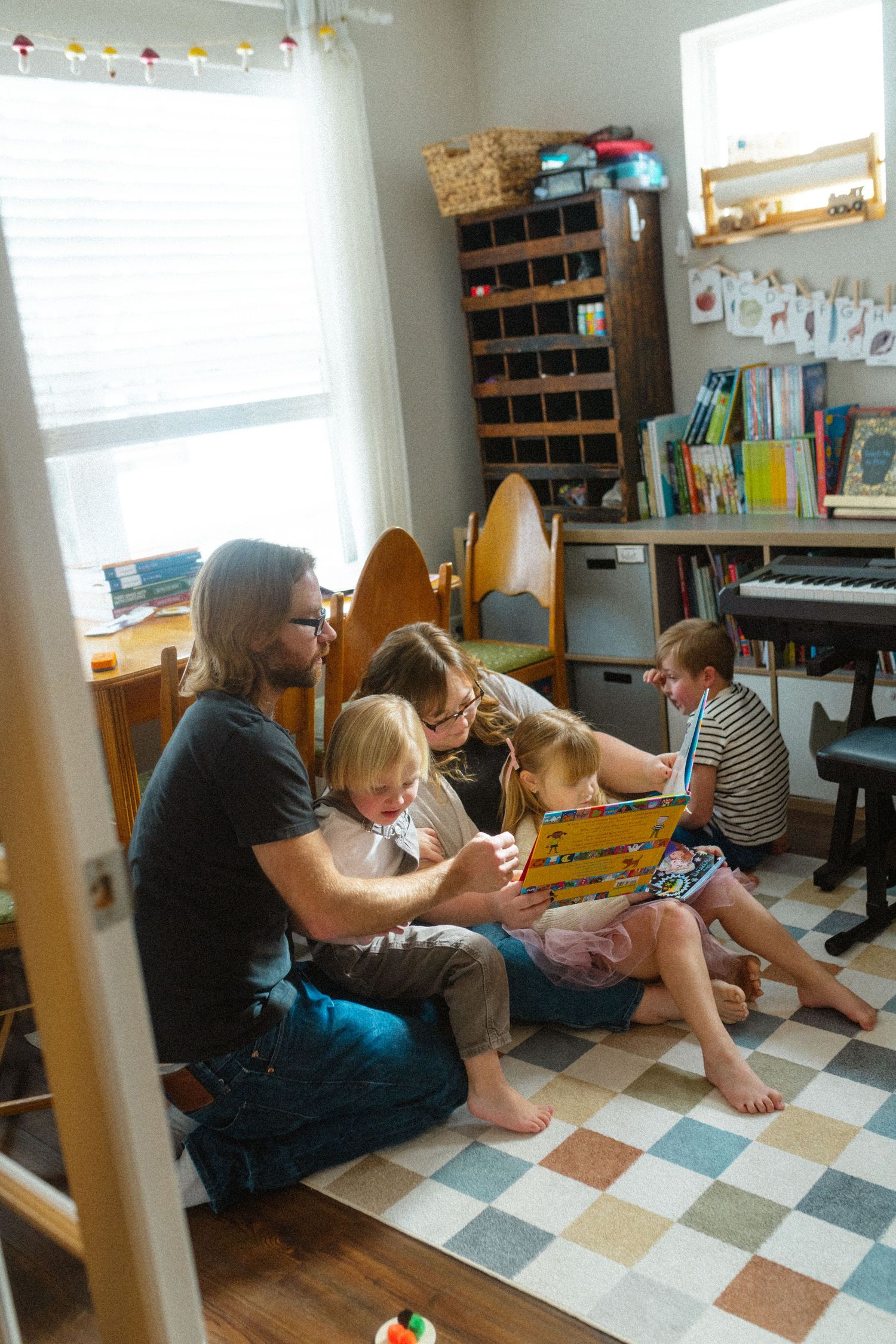 Family sitting on floor reading a colorful book together in a cozy children's playroom with bookshelves.