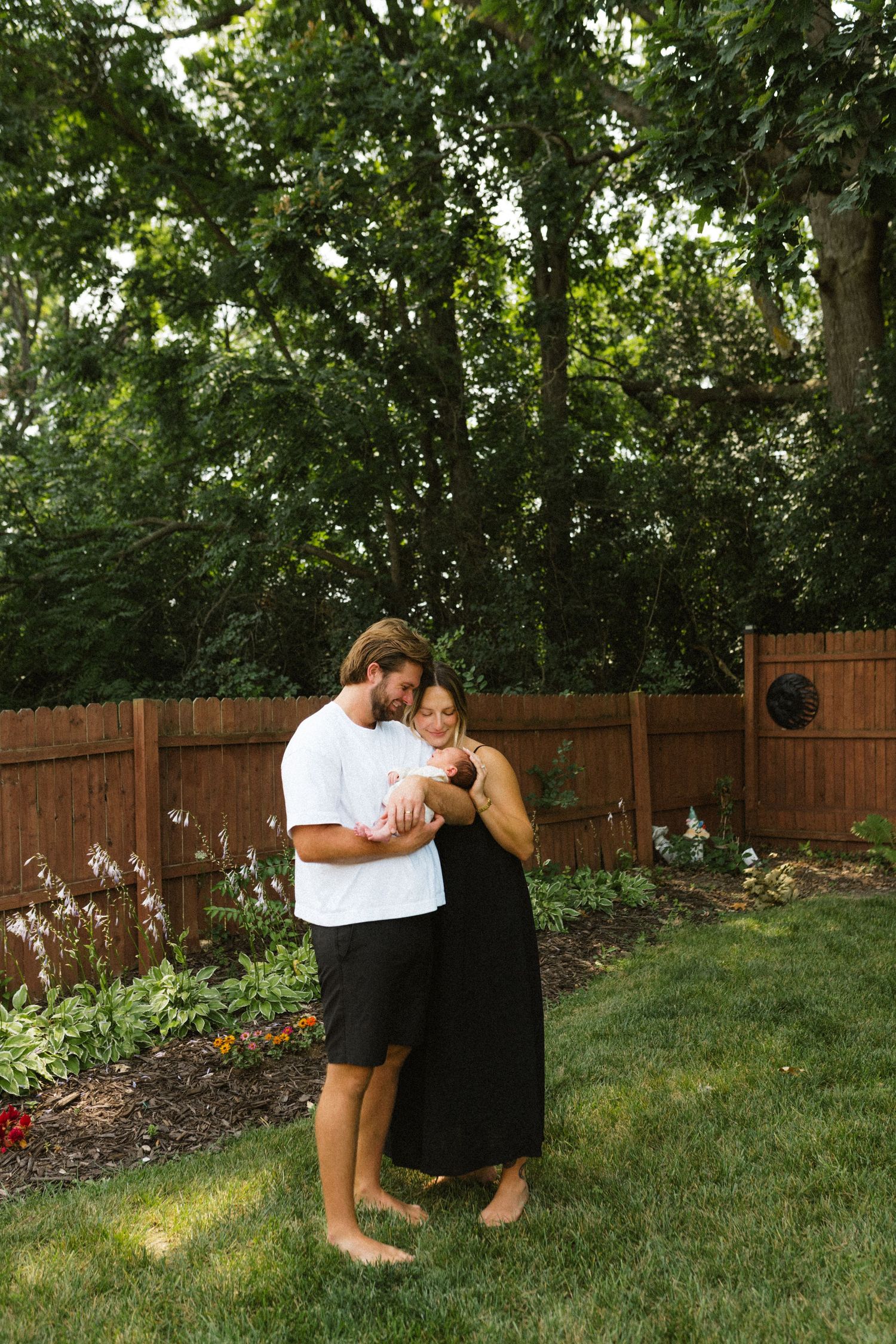 Couple holding newborn baby barefoot in lush green backyard garden near wooden fence.