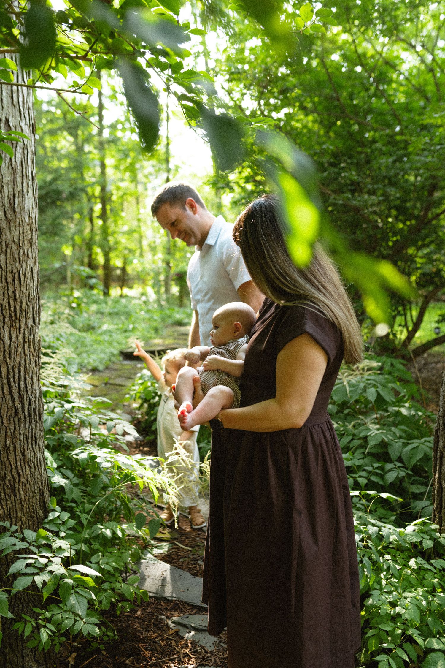 Parents admiring newborn baby on a shaded garden path surrounded by lush green foliage and stepping stones.