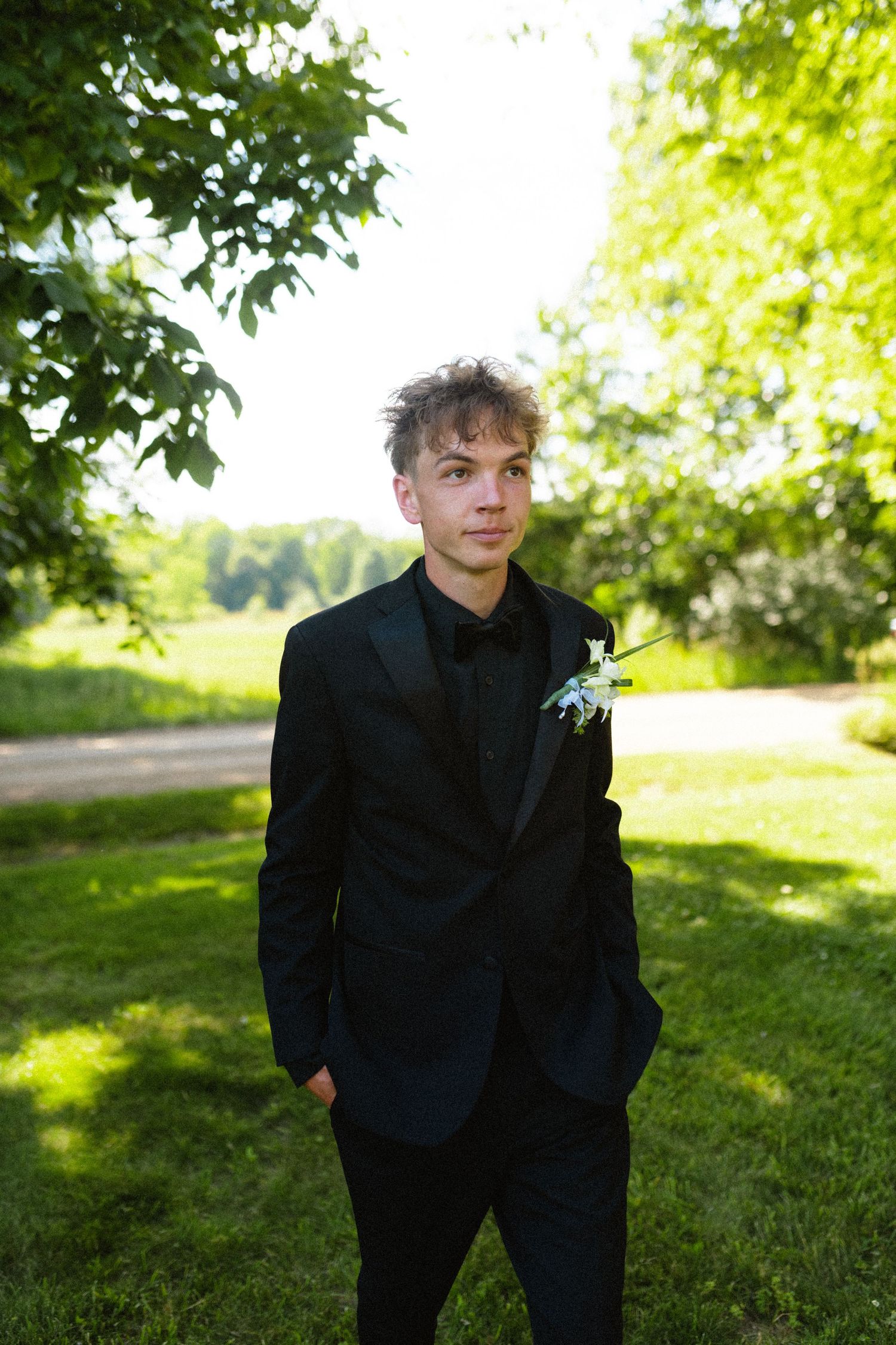 Young groom in black tuxedo with boutonniere stands outdoors.