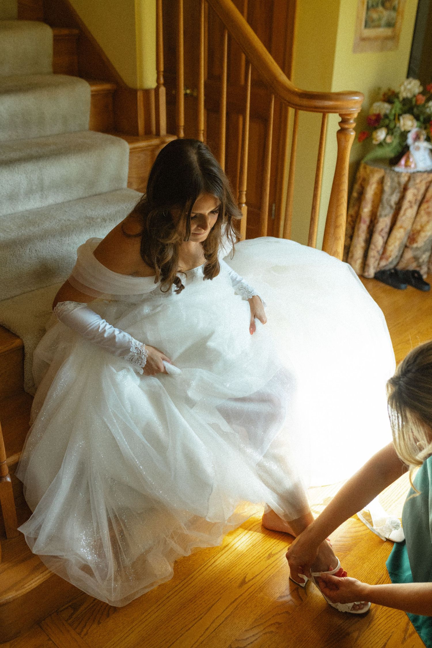Bride in white off-shoulder gown getting shoes fastened by bridesmaid on wooden staircase.
