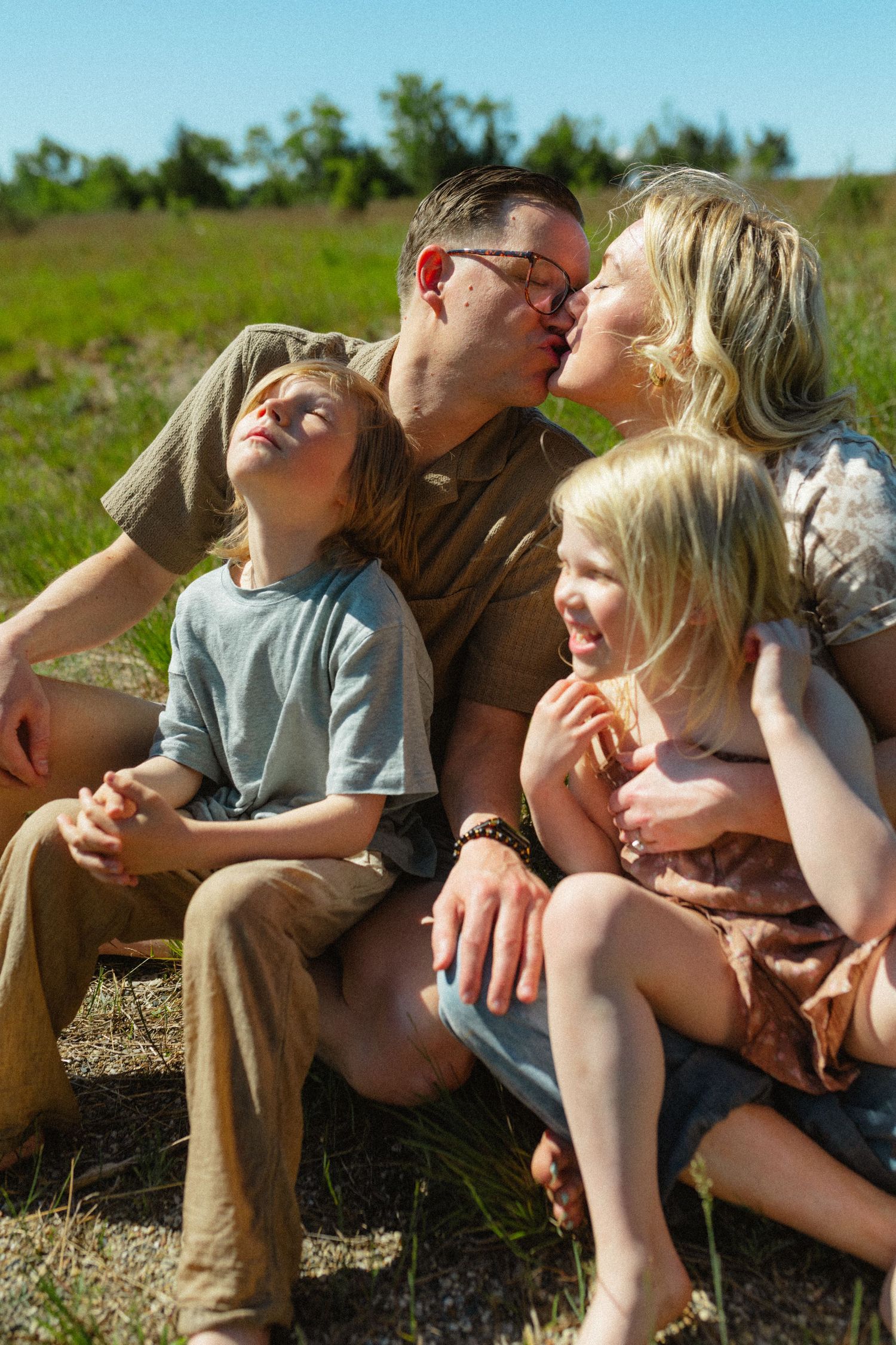 Family of four sitting together in a sunny field as parents share a kiss with two kids.