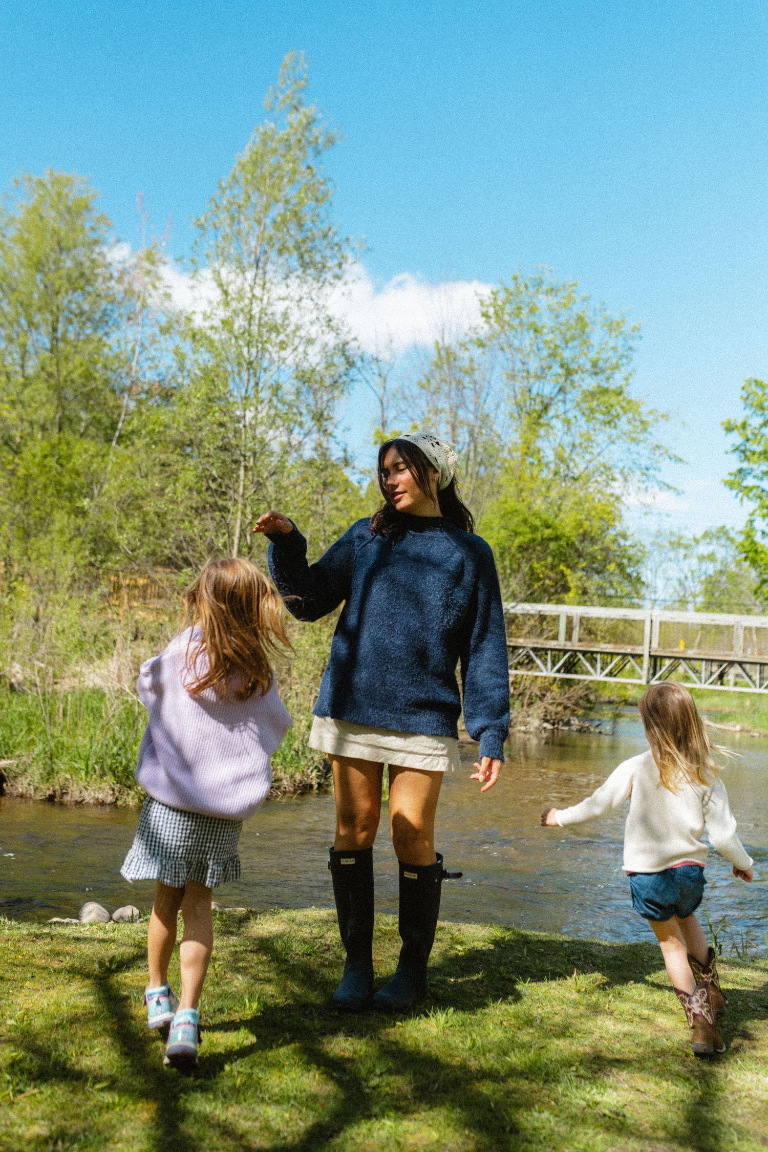 Mother in blue sweater and rain boots playing with two young daughters near a sunny riverside.