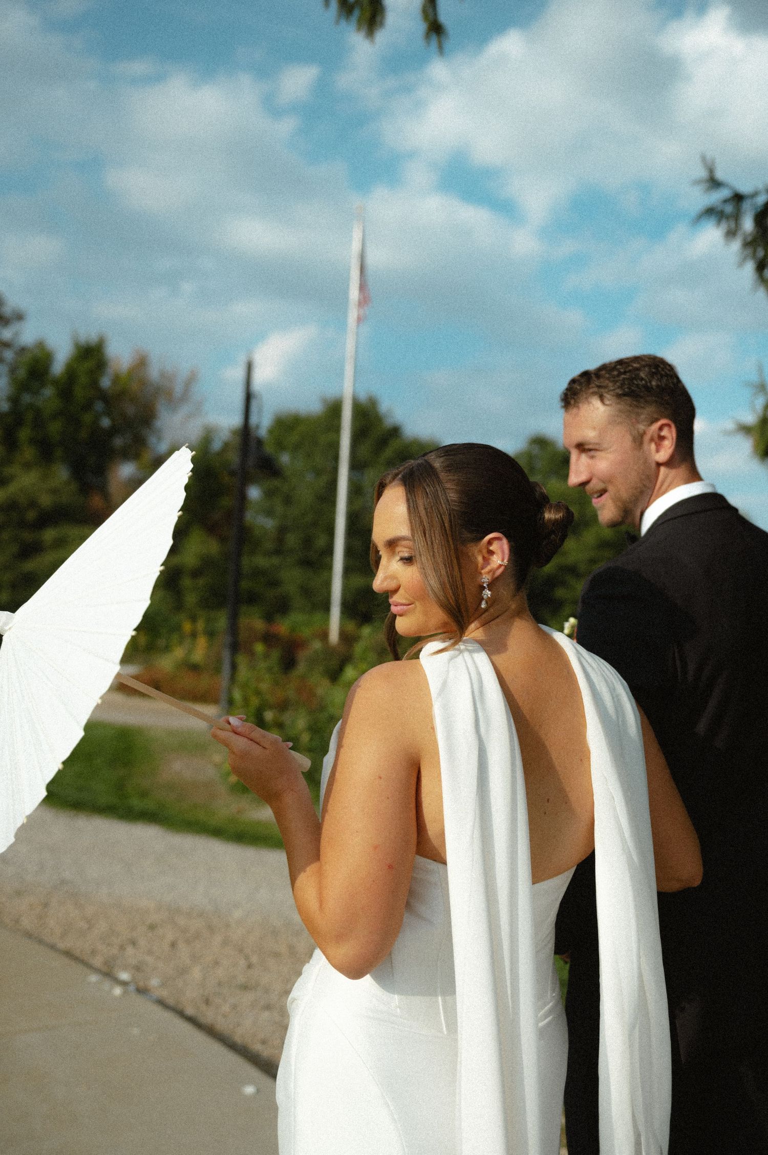 Bride in backless halter gown holding white parasol with groom smiling under blue sky.