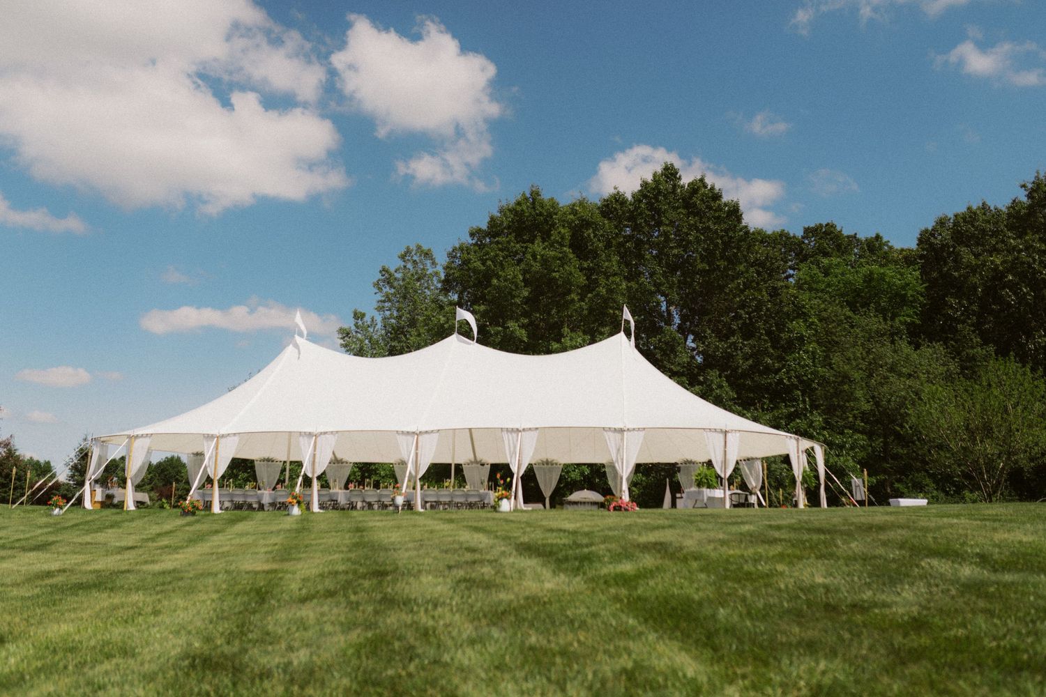 Large white sailcloth wedding tent on green lawn under blue sky with clouds.