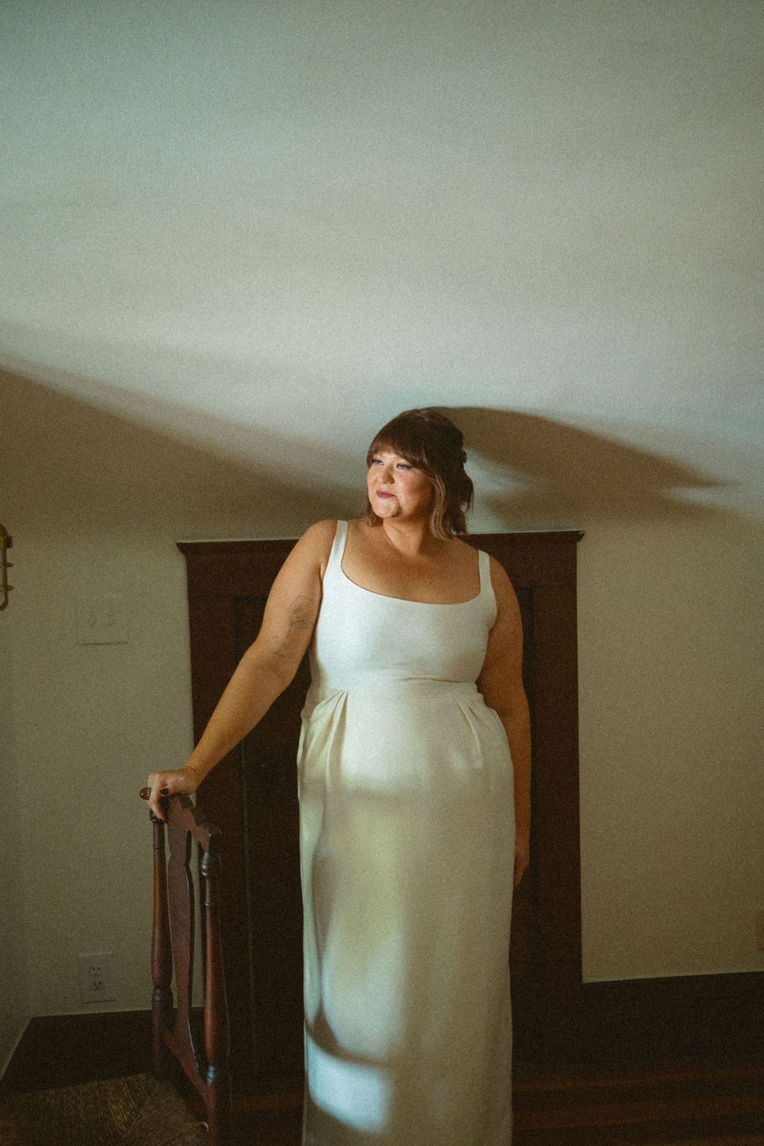 Bride in simple white sleeveless wedding gown posing by wooden chair in softly lit room.