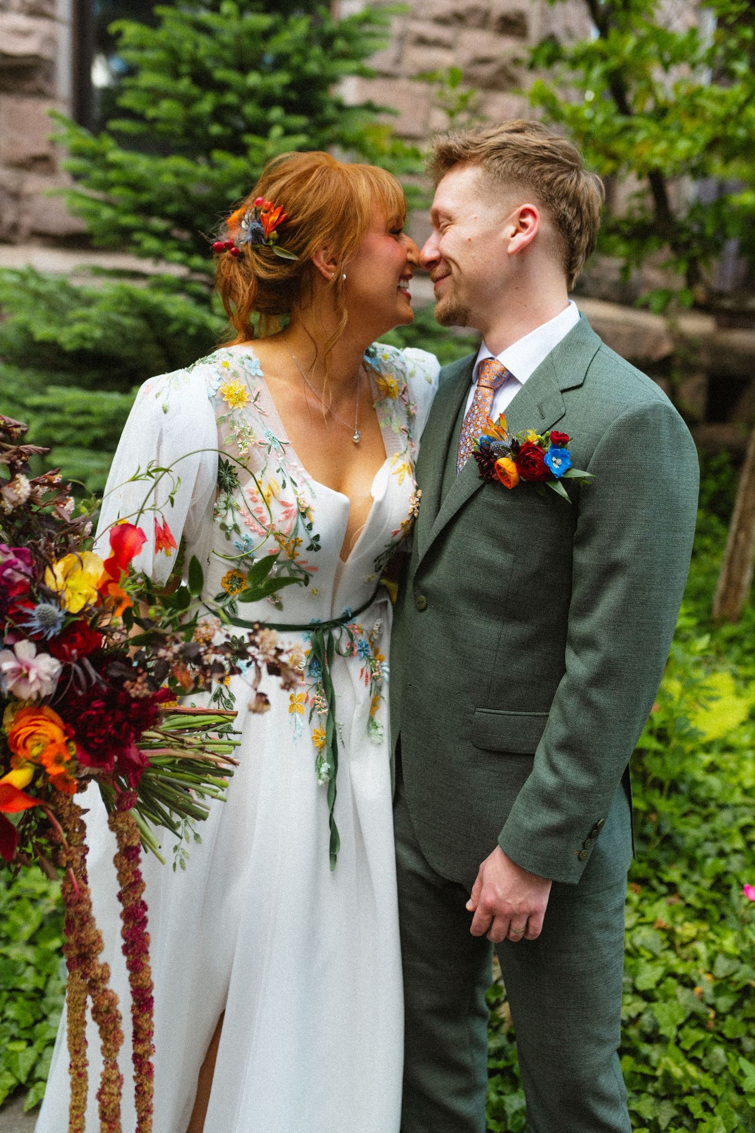 Bride in floral embroidered gown and groom in green suit sharing intimate moment with colorful bouquet.