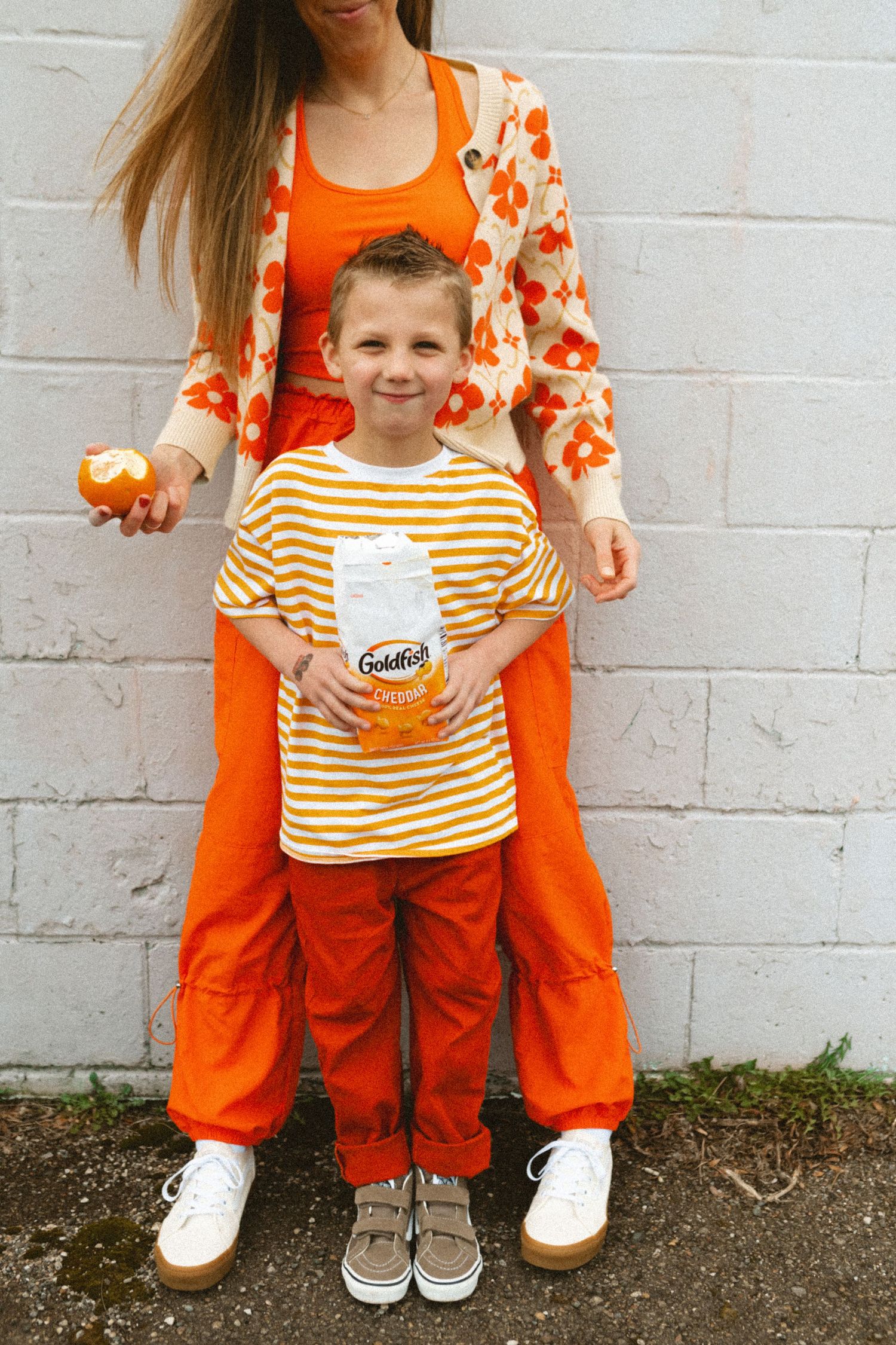 Mother and son in matching orange outfits holding Goldfish crackers against a white brick wall.