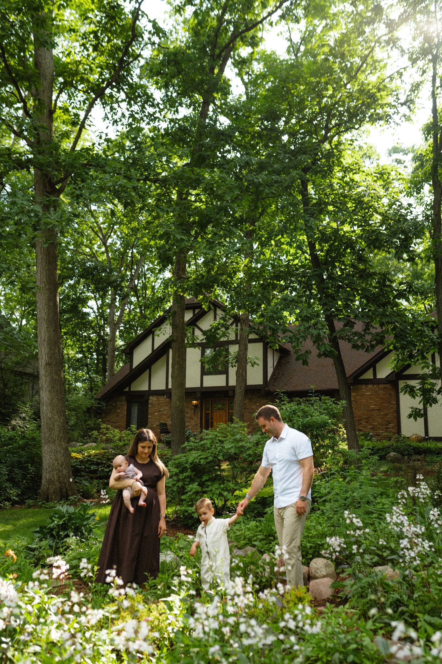 Family of four walking through a lush garden in front of a Tudor-style home surrounded by trees.
