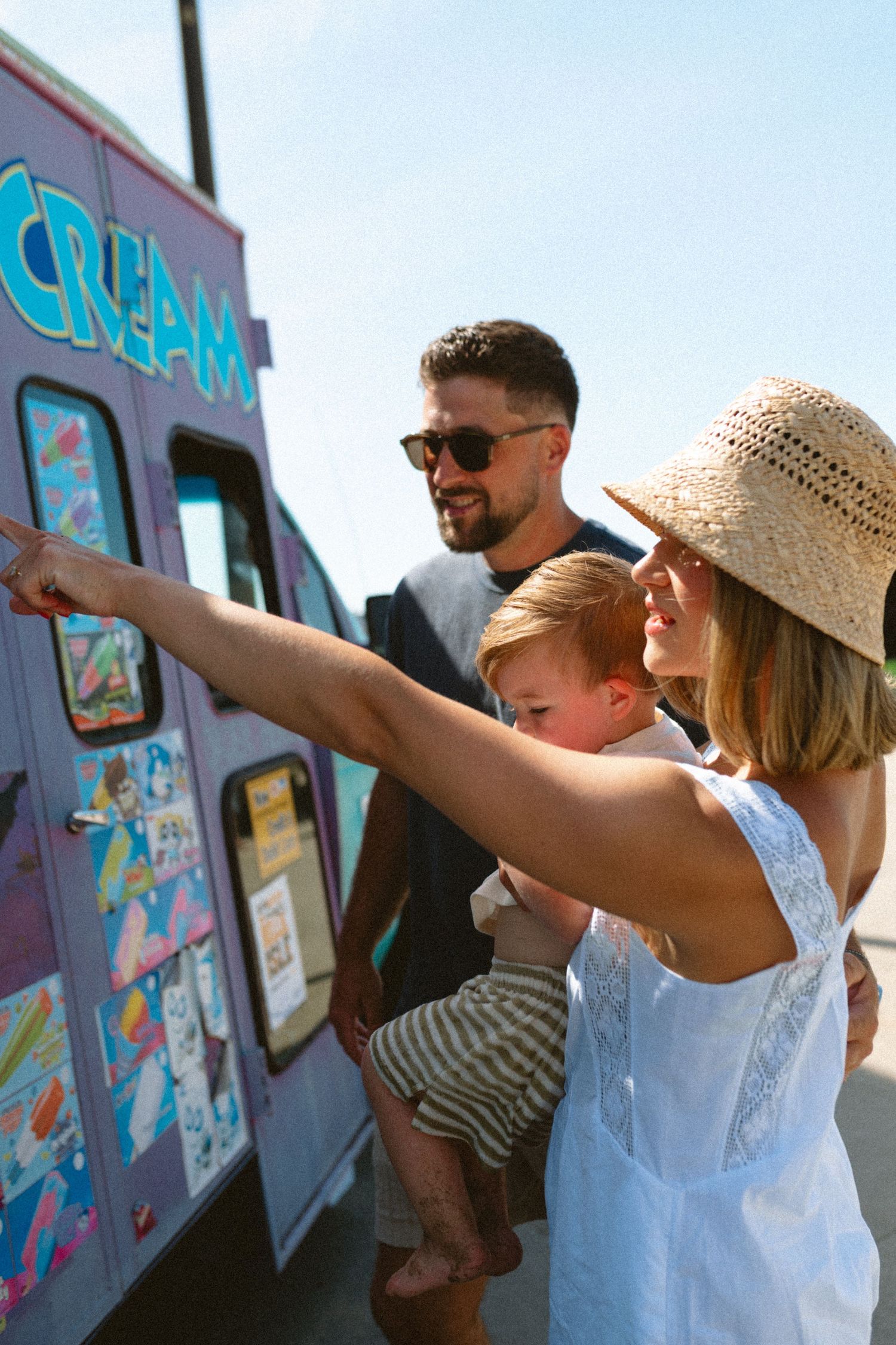 Mother pointing at ice cream truck menu while holding toddler son with smiling father behind.