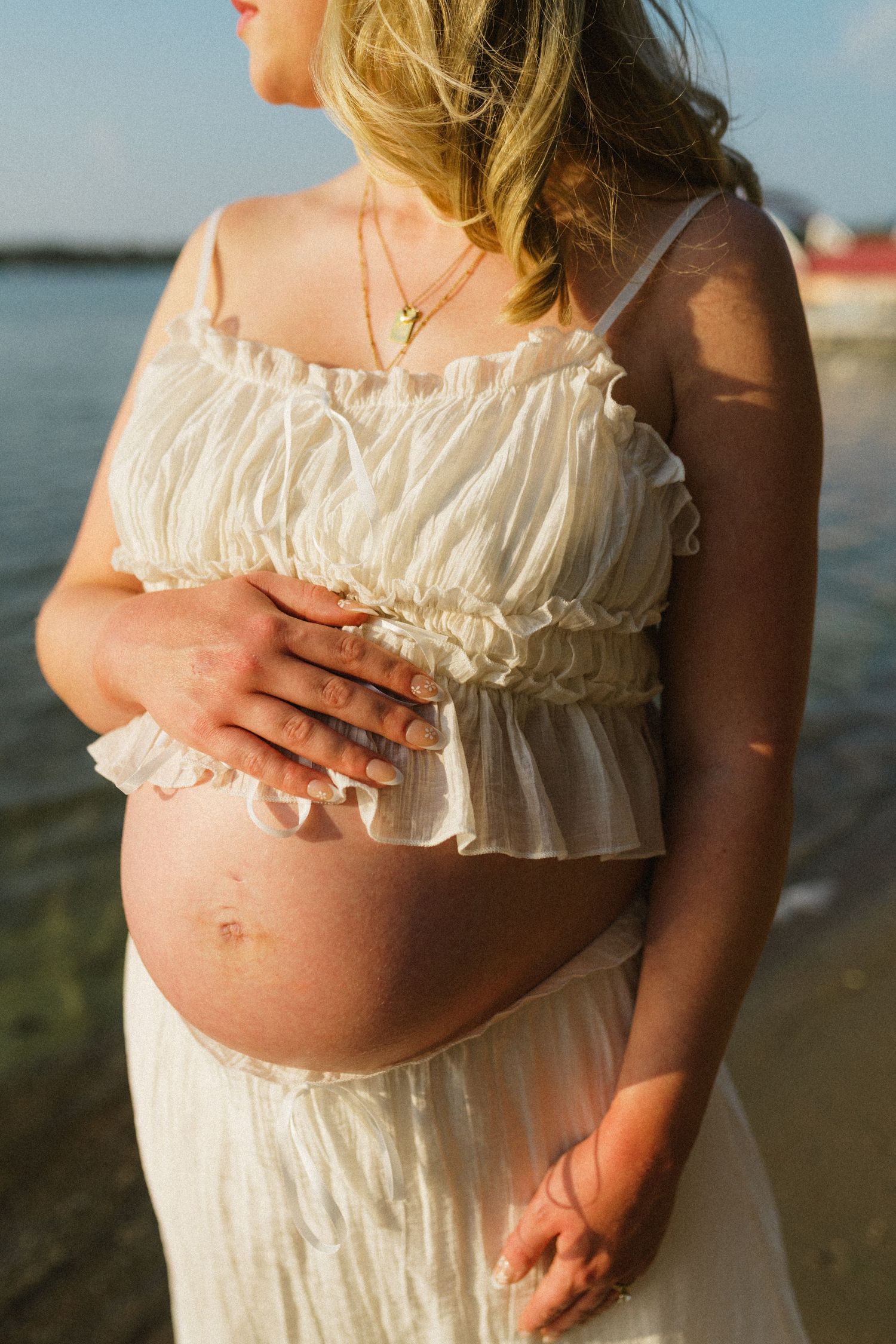 Close-up of a pregnant woman in a white ruffled crop top cradling her bare baby bump outdoors.