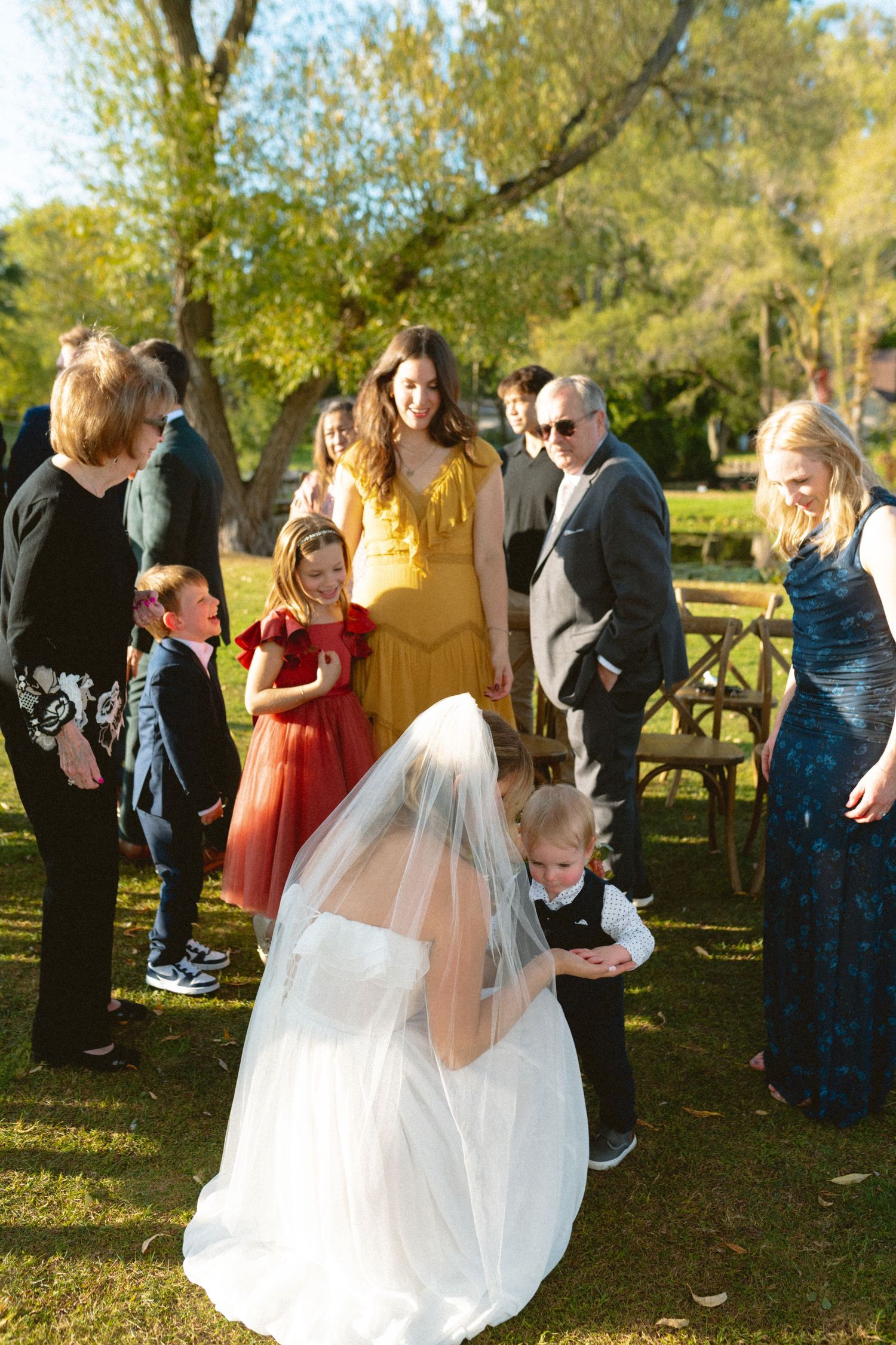 Bride in white gown and veil kneels to greet young children at outdoor wedding reception.
