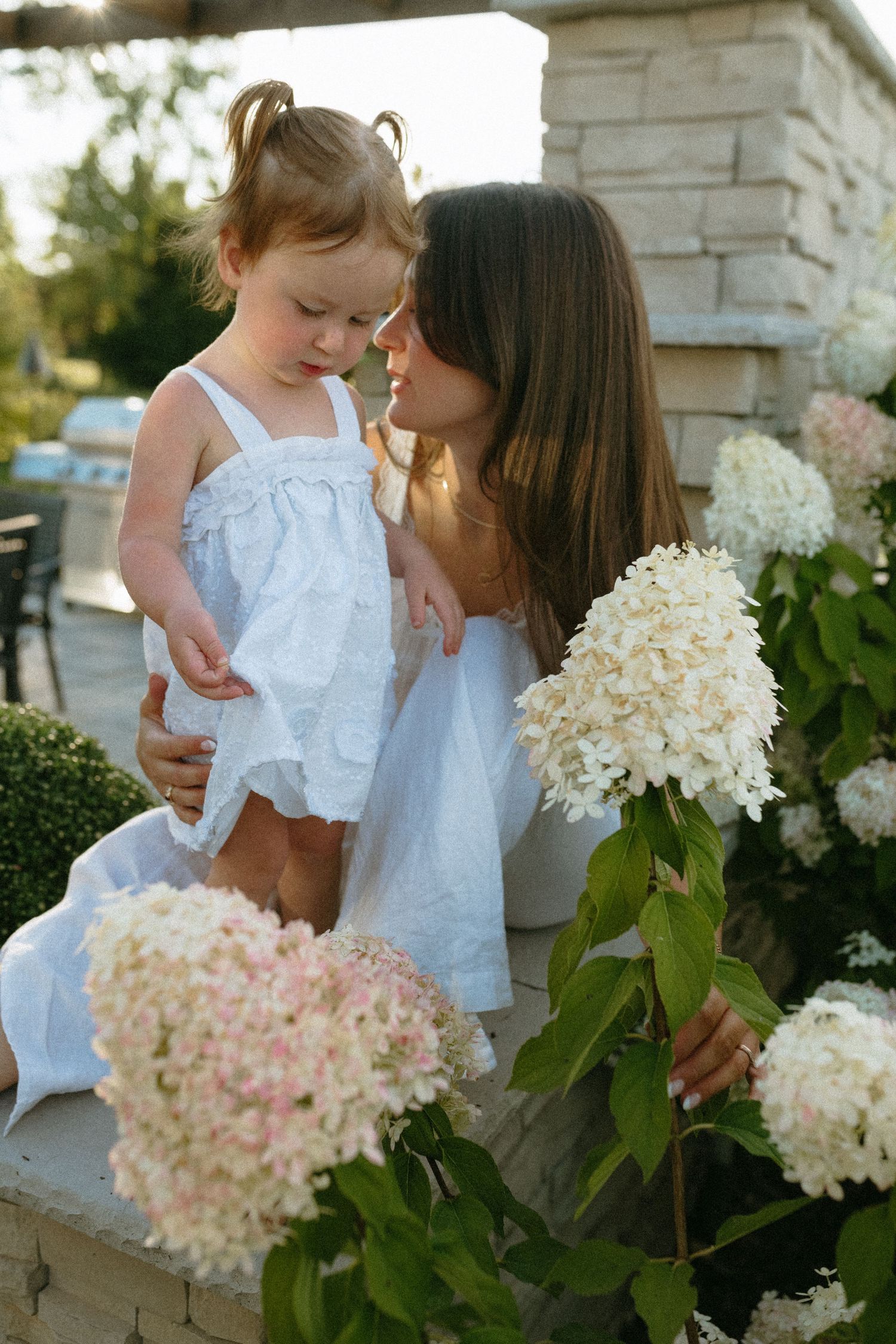 A mother in white tenderly holds her toddler daughter near blooming white hydrangeas outdoors.
