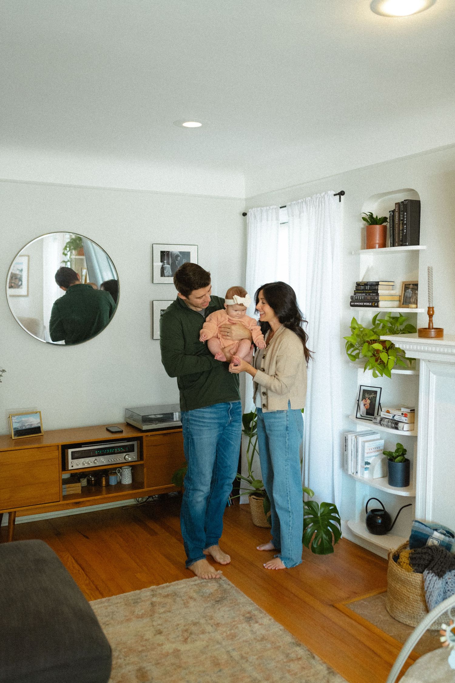 Young couple admiring their newborn baby in a bright, modern living room with shelves and plants.