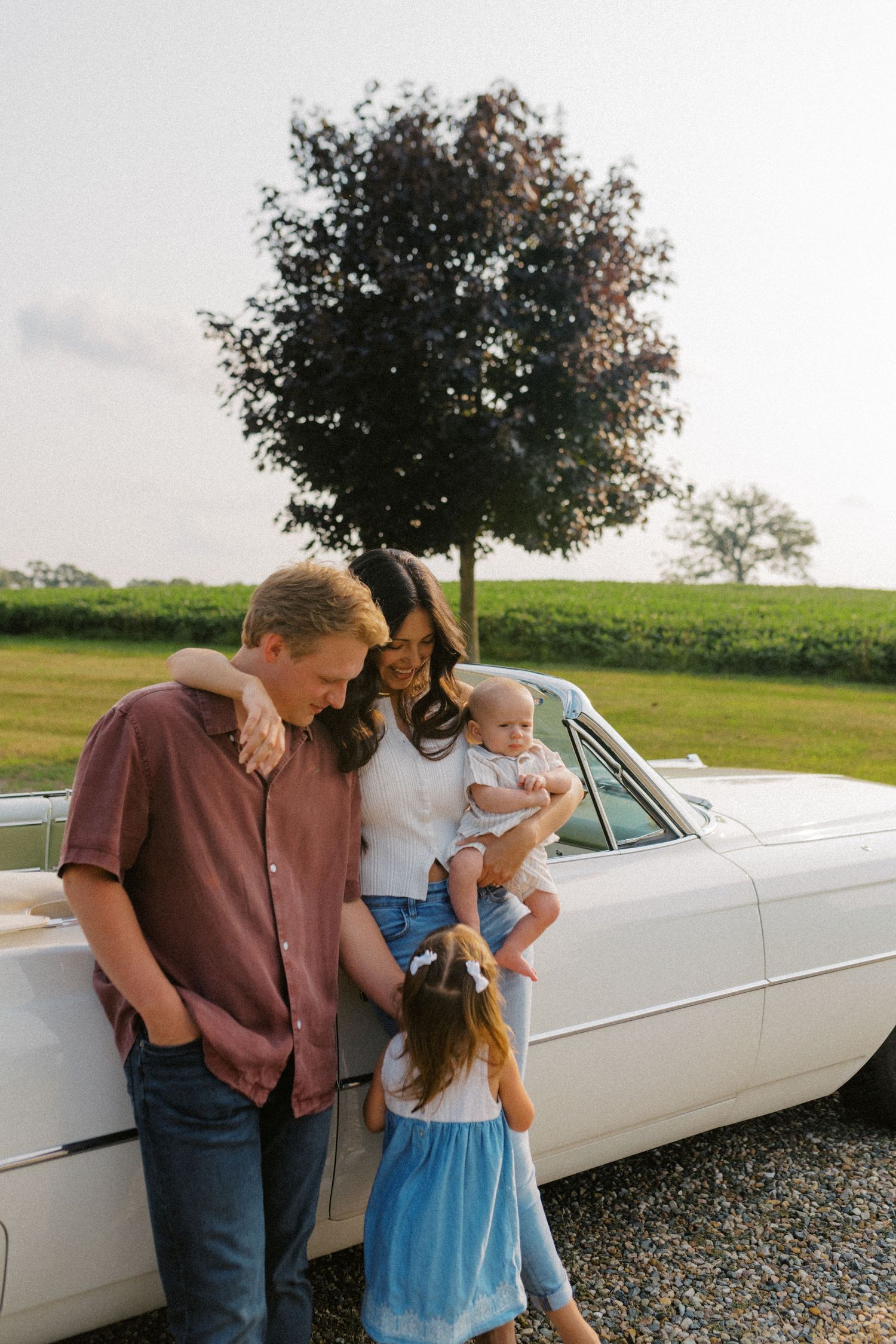 Family of four with baby and toddler posing beside a vintage white car in a rural setting.