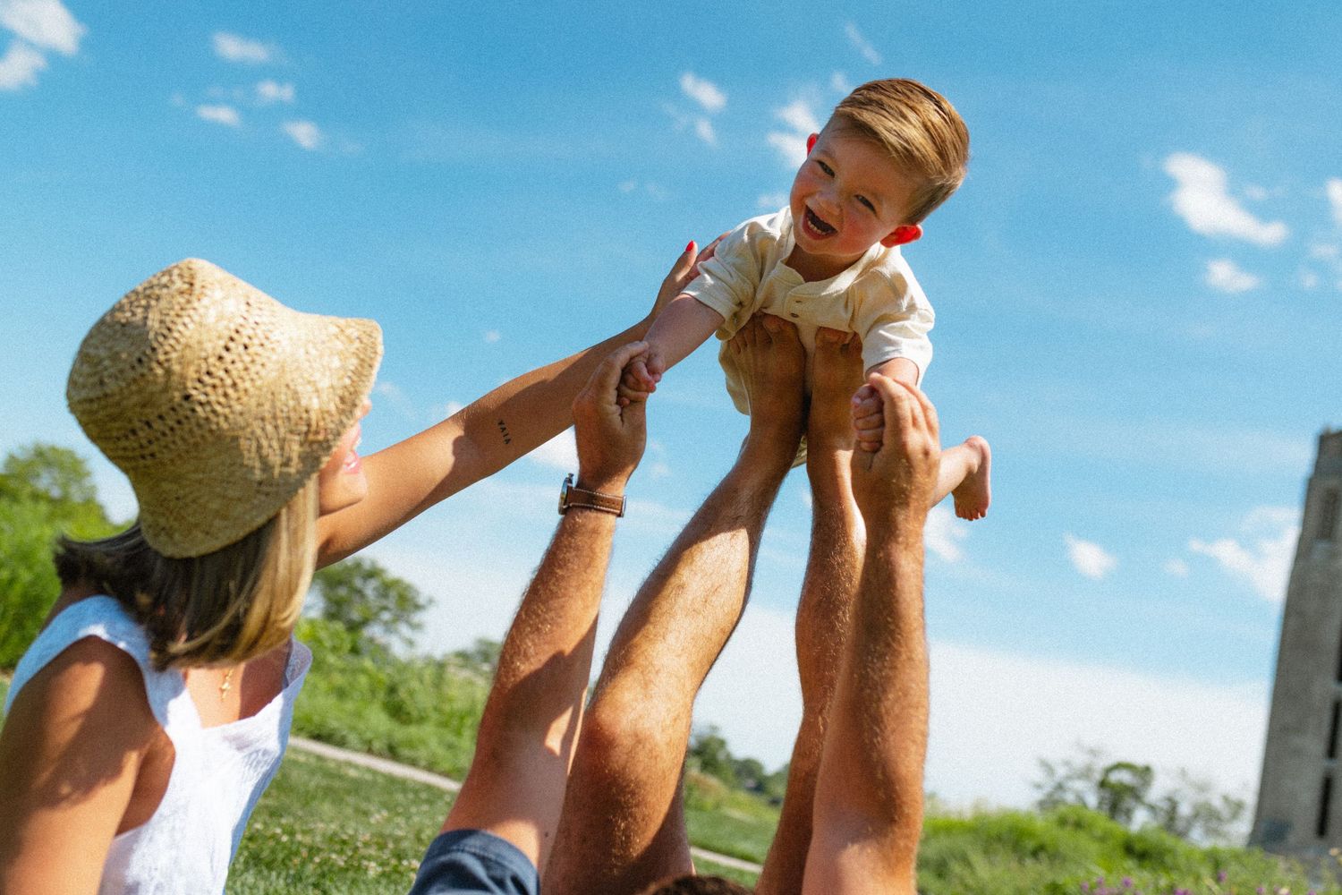 Happy parents lifting laughing toddler boy high in the air against a bright blue sky in a green park.