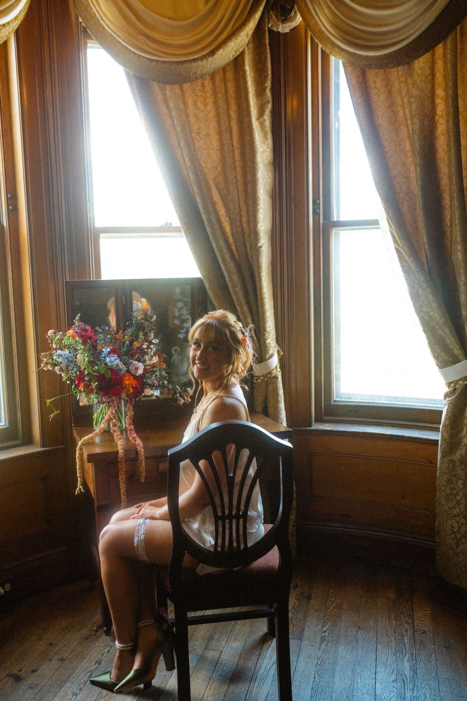 Bride in lingerie sits on chair by sunlit vintage draped windows.