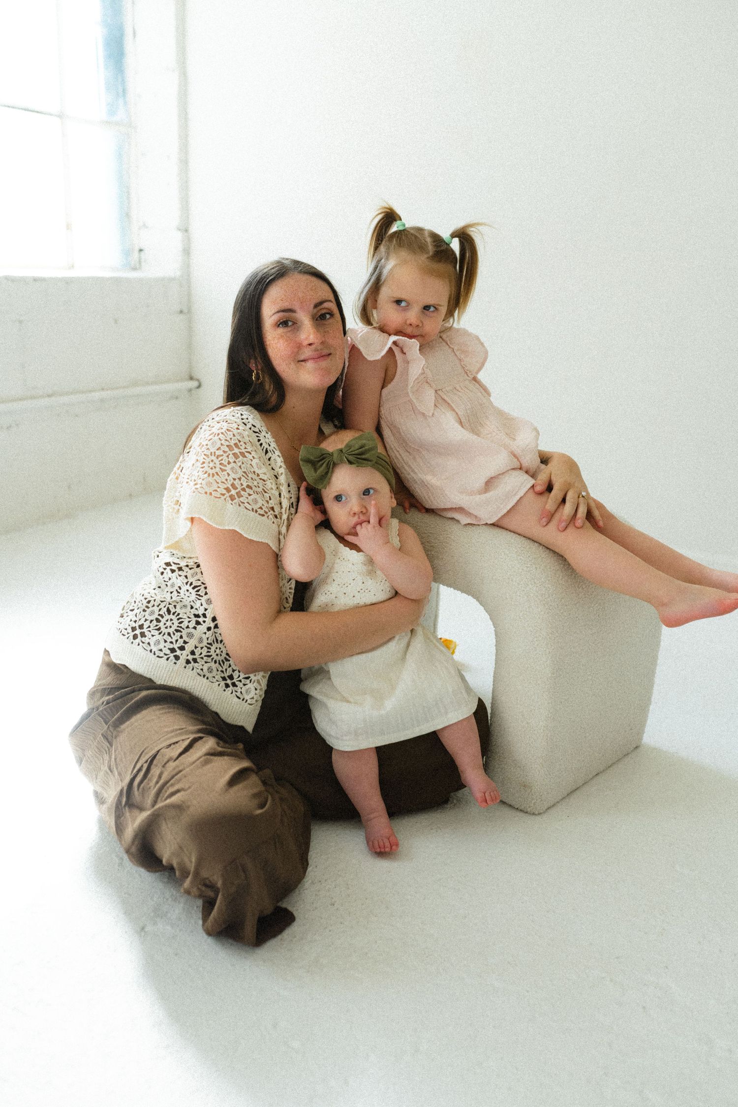 A mother sits on the floor with two young daughters, one baby and one toddler, in a bright white studio setting.