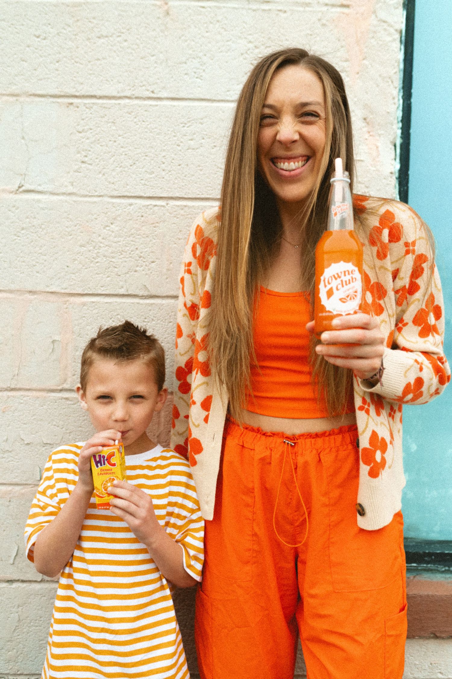 Smiling woman in orange outfit holding Town Club soda bottle with boy drinking Hi-C.
